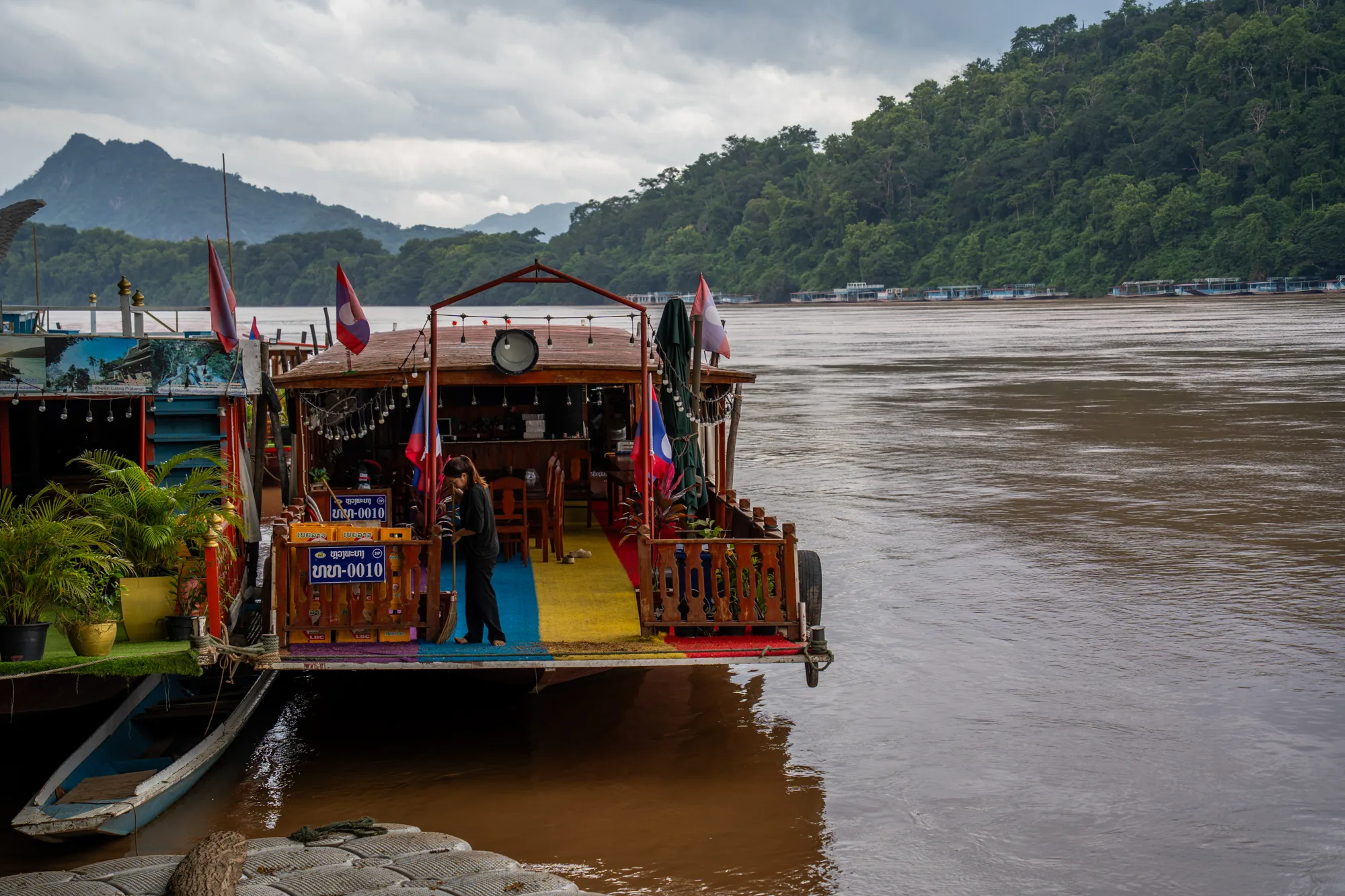 この写真はラオスのルアンパバーンで撮影された風景を捉えています。前景には川に浮かぶ船が写っており、その船はレストランかバーのような施設として使われているようです。船尾にはラオスの国旗が掲げられ、装飾的なライトが飾られています。船のデッキは赤、黄、青の鮮やかな色で塗られています。  船のデッキでは女性がほうきで掃除をしています。傍らにはラオス語で書かれたナンバープレートのようなものが見えます。船の脇には小さなボートが係留されており、植木鉢に入った植物が置かれています。  背景には茶色く濁った川の流れが広がり、対岸には緑豊かな森林が広がっています。遠くには山々が霞んで見え、空は曇っています。全体的に落ち着いた雰囲気で、東南アジアののんびりとした空気感が伝わってきます。川の色から、雨季か洪水後であることが伺えます。