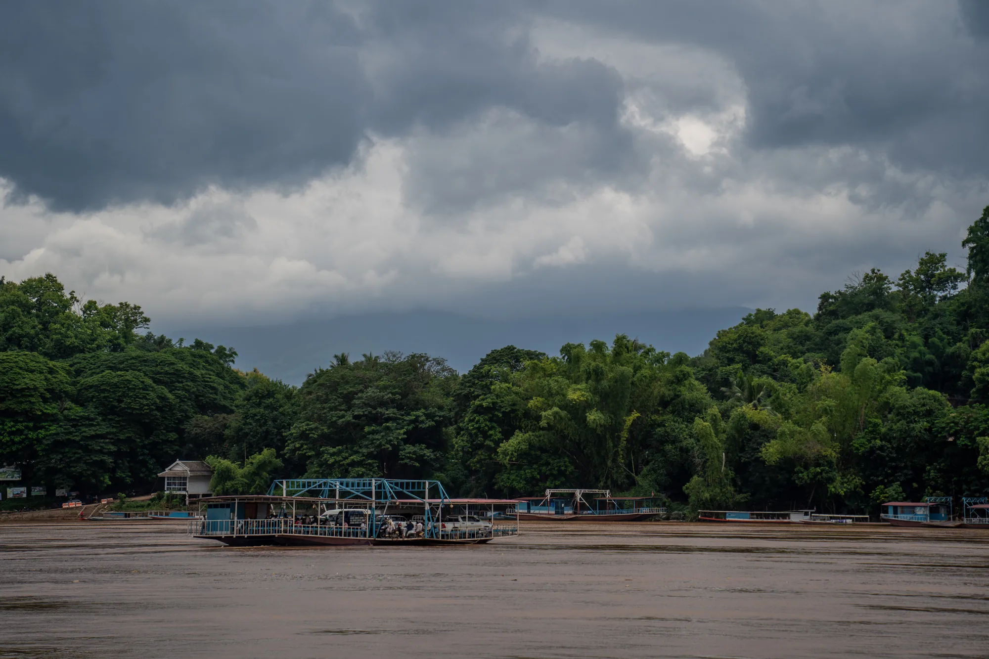 ラオスのルアンパバーンで撮影された写真です。画面中央には、メコン川を渡るための、青い鉄骨で組まれた構造の渡し舟が写っています。この船には車や人々が乗っており、対岸へ向かっているようです。川の水は茶色く濁っており、雨季の増水期であることが伺えます。対岸の岸辺には、緑豊かな熱帯の木々が生い茂っています。奥には、かすかに山並みが見えます。空は厚い雲に覆われており、今にも雨が降り出しそうな、重苦しい雰囲気です。全体的に落ち着いた色調で、静寂と湿気を感じさせる写真です。渡し舟は人々の生活に欠かせない交通手段であることが伝わってきます。
