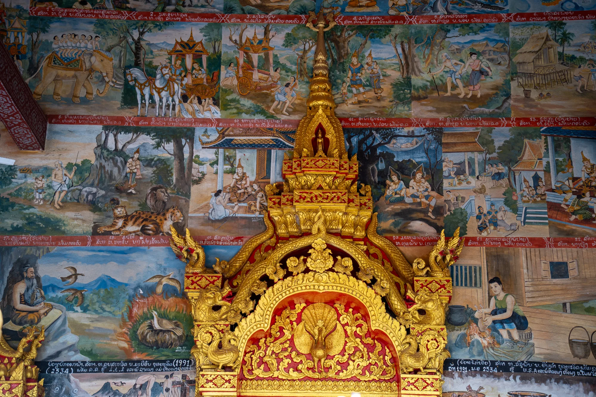 この写真は、ラオスのルアンパバーンで撮影された寺院の一部を捉えたものです。画面中央には、鮮やかな赤と金を基調とした装飾的な仏塔の一部が大きく写っています。この仏塔は複雑な彫刻が施されており、孔雀のような鳥のモチーフや、その他の装飾的な要素が目を引きます。  仏塔の背景には、壁一面に描かれた壁画が広がっています。壁画は複数の場面に区切られており、それぞれが異なる物語を描いているようです。象に乗った人々、馬車、風景、人々が生活する様子などが、鮮やかな色彩で細かく描かれています。全体的に歴史的な物語や伝説を伝えるような、宗教的または文化的な雰囲気を感じさせます。また、壁画にはラオス語の文字と思われるテキストも含まれています。  壁画のタッチや色使いは、どこか素朴で味わい深く、その歴史を感じさせます。写真全体の構成は、中央の仏塔と背景の壁画が組み合わさることで、独特な奥行きと視覚的な面白さを生み出しています。寺院の歴史、文化、そして芸術性が凝縮された一枚と言えるでしょう。