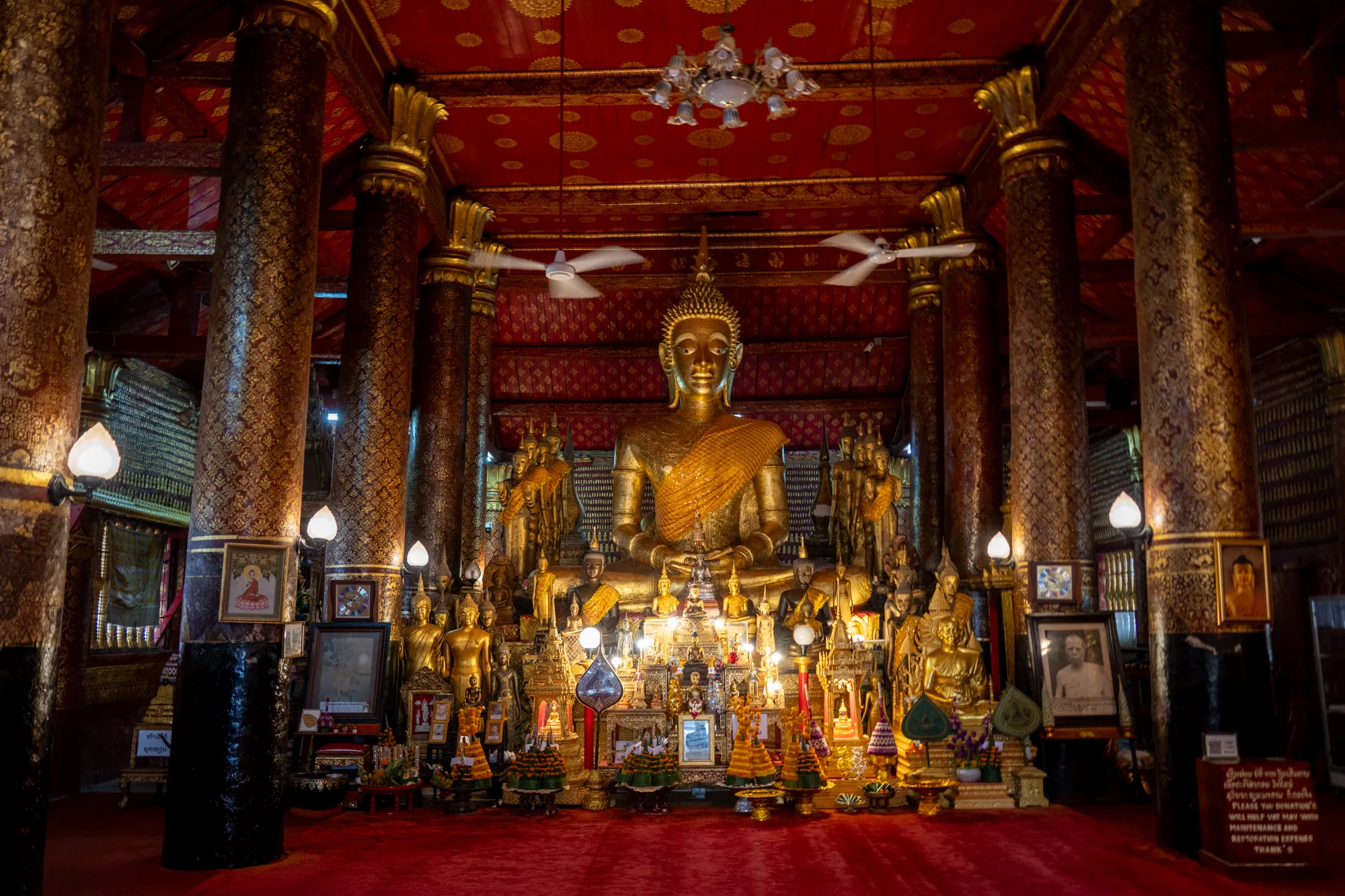 ラオスのルアンパバーンにある寺院内部を写した写真です。メインには金色に輝く大きな仏像が鎮座しており、その周りにはさらに多くの仏像や装飾品が並べられています。床は鮮やかな赤色の絨毯で覆われており、天井は赤色と金色で装飾され、シャンデリアやシーリングファンが設置されています。柱は金色模様が施されており、寺院全体の豪華さを際立たせています。雰囲気は神聖で静謐。左右には写真や寄贈のお願いの看板などが見られます。