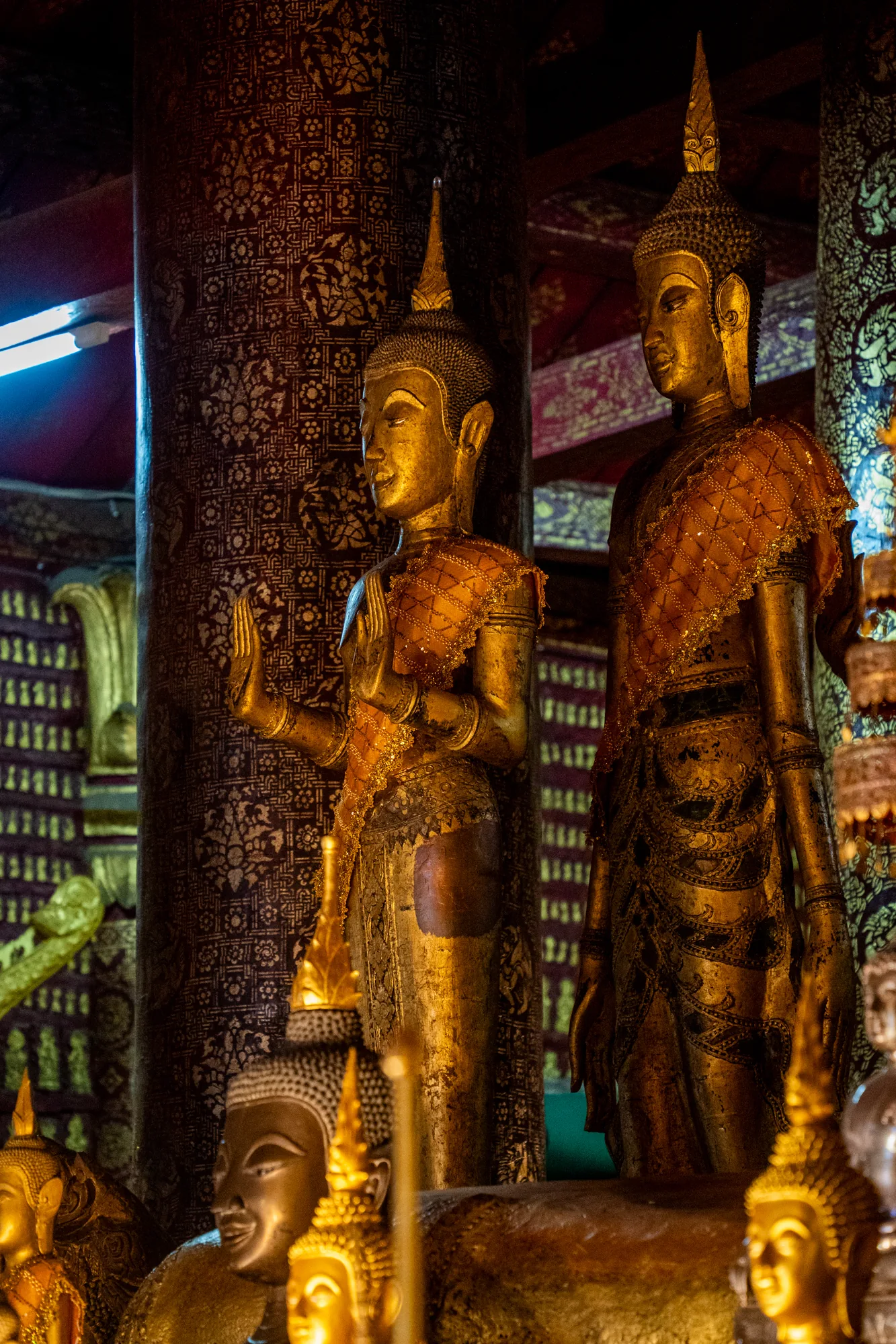 ラオスのルアンパバーンで撮影された写真です。写真は仏像が複数安置されている様子を捉えています。主な被写体は、金色の美しい仏像です。手前に小さめの仏像の頭部が複数あり、奥には直立した二体の仏像が立っています。手前の仏像の顔はにこやかに微笑んでいるように見えます。直立した仏像は、繊細な装飾が施された衣装を身に着けています。一体は右手を上げ、もう一体は静かに佇んでいます。背景には模様が施された柱が立ち並び、照明の光が当たっています。全体的に落ち着いた雰囲気で、仏教の神聖な空間を表現しているようです。