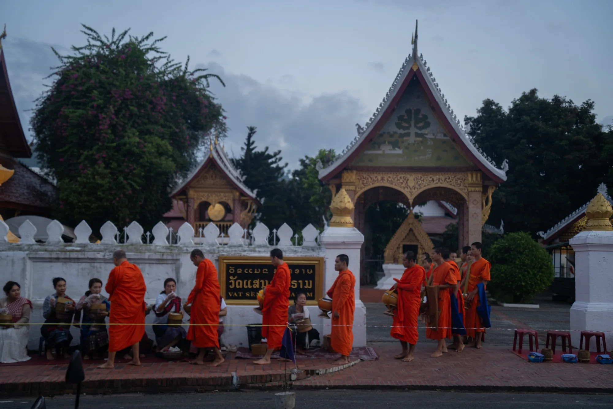 写真はラオスのルアンパバーンで撮影されたもので、早朝の托鉢の様子を捉えています。背景には伝統的な寺院の建物が見え、金色の装飾が施された屋根が特徴的です。空は薄暗く、まだ夜明け前の時間帯であることが分かります。手前には、鮮やかなオレンジ色の袈裟をまとった僧侶たちが、お供え物を受け取るために列を作って歩いています。人々は僧侶たちに敬意を払いながら、食べ物や飲み物などを施しています。写真全体から、静かで神聖な雰囲気が感じられます。ルアンパバーンの文化的な側面を捉えた一枚と言えるでしょう。