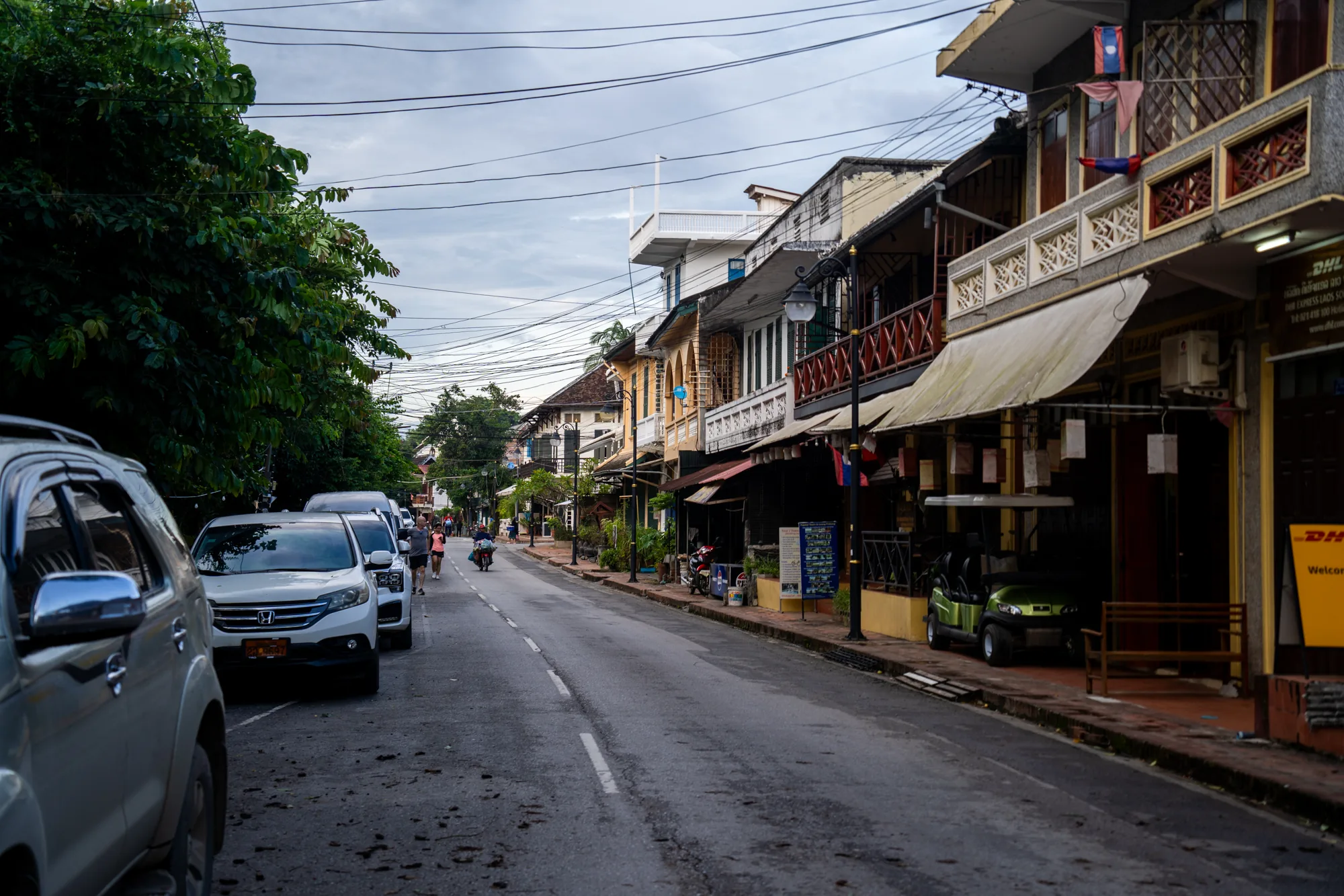 ラオスのルアンパバーンで撮影されたと思われる写真です。アスファルトの道路が画面中央を走り、その左側には車が数台縦列駐車しています。道路の右側には、歴史を感じさせる建物が並んでいます。建物は2階建て、もしくは3階建てで、バルコニーが付いているものもあります。建物の多くは店舗や宿泊施設のようです。軒先には日よけのオーニングが出ており、ラオスの国旗が飾られている建物も見られます。道路を歩く人やバイクに乗る人の姿も見え、のどかな街の雰囲気が伝わってきます。電線が空を覆い、曇り空であることから、天候はあまり良くないことが伺えます。全体的に落ち着いた色調で、静かで穏やかな時間が流れているような印象を受けます。