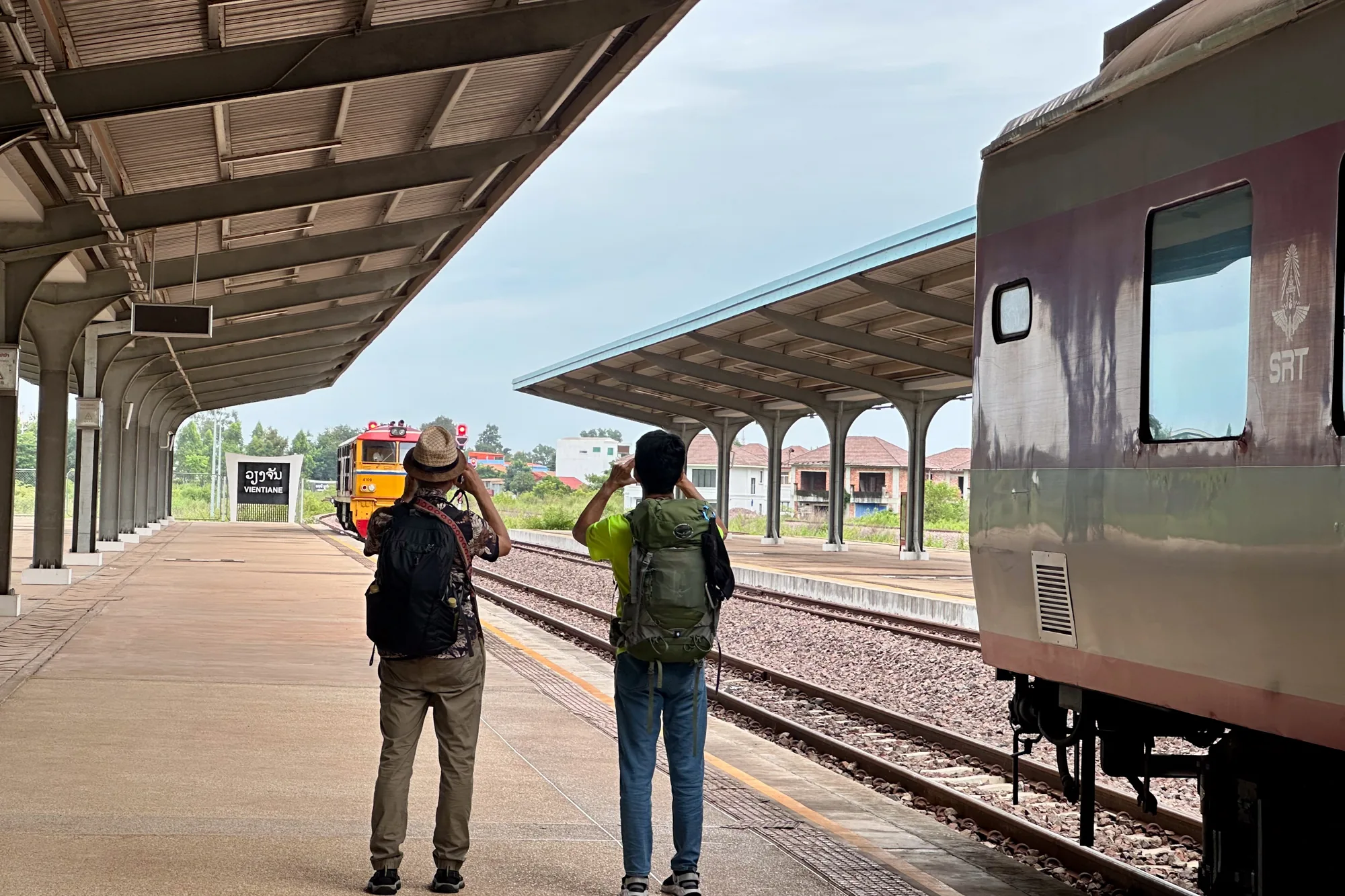 この写真はラオスのビエンチャンで撮影された、駅のプラットフォームの風景です。手前にはプラットフォームに立っている二人の男性の後ろ姿が写っています。左側の男性は帽子をかぶり、花柄のシャツにズボン姿で、大きなバックパックを背負っています。右側の男性は黒い帽子をかぶり、蛍光色の黄緑色のシャツにジーンズ姿で、緑色のバックパックを背負っています。二人は線路の奥の方を見ており、何かを眺めているようです。  線路の向こう側には「VIENTIANE」と書かれた駅の看板があり、その奥にはオレンジ色の機関車が停車しています。また、右側のプラットフォームには列車の一部が写っており、車体には「SRT」のロゴが見えます。  プラットフォームの上には、屋根が設置されており、日差しを遮っています。全体的に曇り空で、落ち着いた雰囲気の写真です。