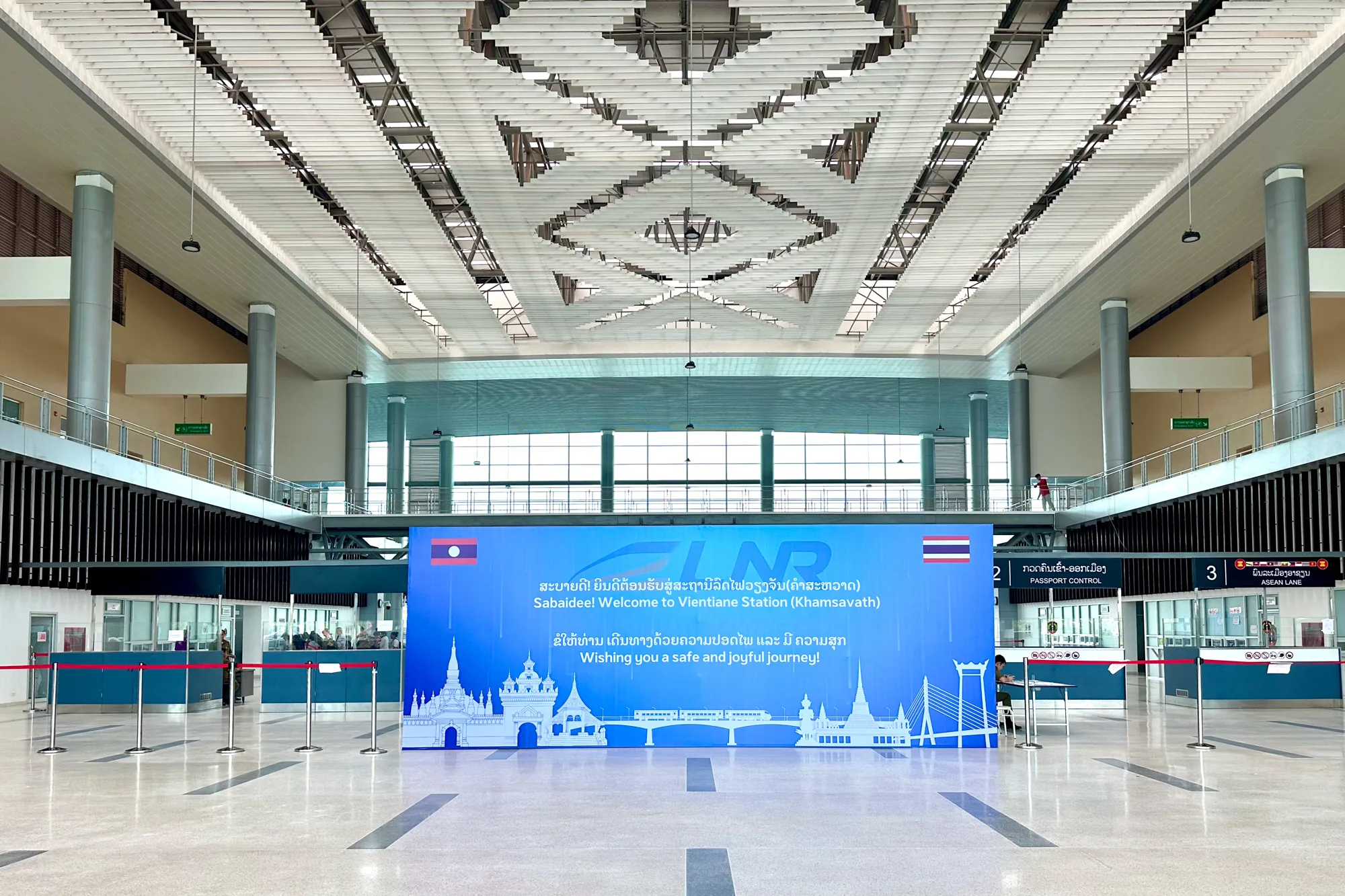 写真は、ラオスのビエンチャンの駅の内部を撮影したものです。広々とした空間で、天井は幾何学的な模様が施された白っぽいデザインで、明るく照らされています。画面中央には、ラオスとタイの国旗が描かれた青い看板があり、「Sabaidee! Welcome to Vientiane Station (Khamsavath)」と英語とラオスの言葉で書かれています。看板の下には、ラオスの寺院や橋などのシルエットが描かれており、駅のデザインを豊かにしています。  手前には、パスポートコントロールのブースがあり、赤いロープで仕切られています。奥には窓があり、いくつかの人が並んでいる様子が見えます。全体的に、駅は清潔で近代的であり、旅行者を歓迎する雰囲気が感じられます。