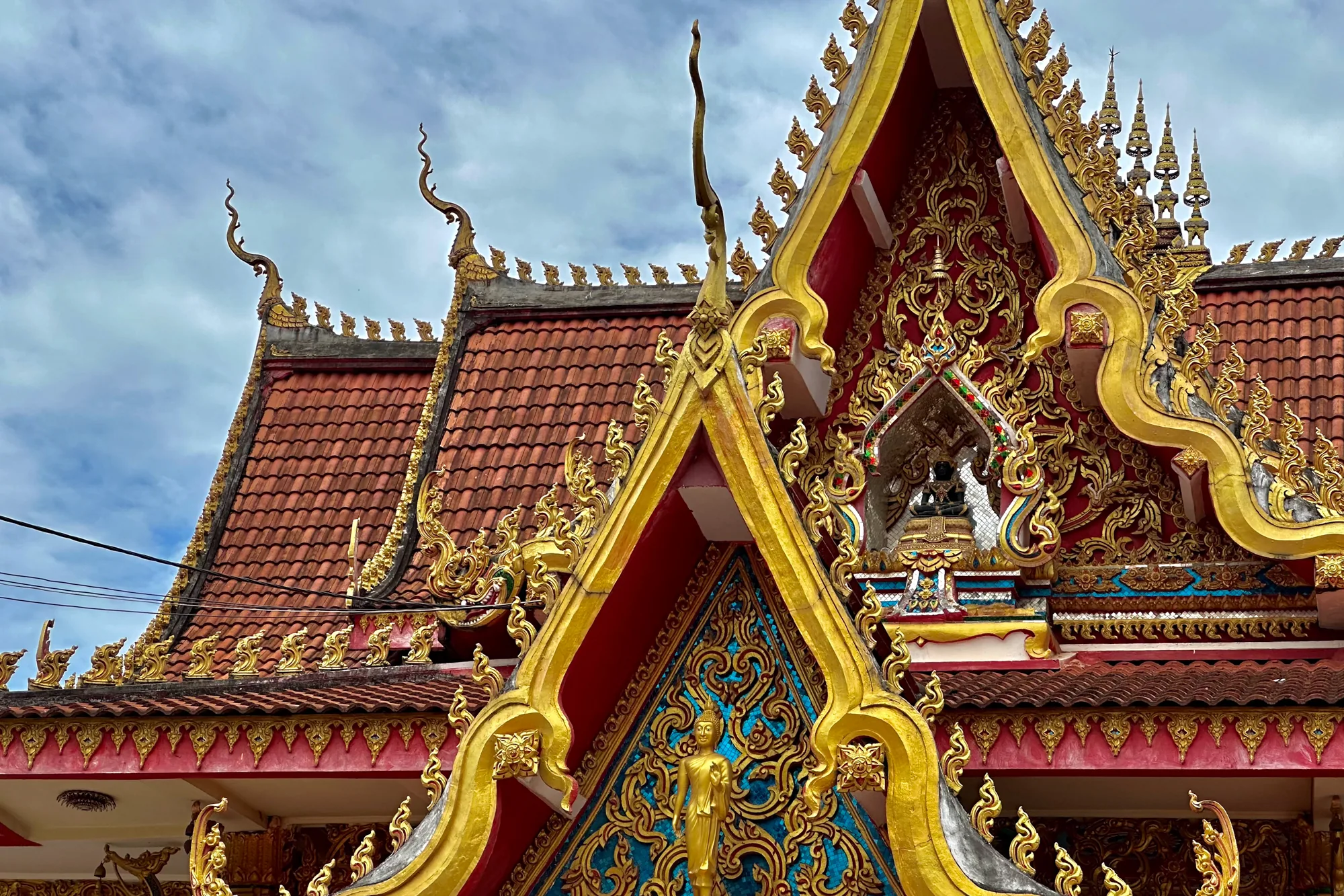 写真は、ラオスの首都ヴィエンチャンにある寺院の建築物の一部を捉えています。画面全体に、鮮やかな赤色の瓦屋根と、金色に輝く複雑な装飾が施された建物が写っています。屋根の縁や軒下には、細部にまでこだわった彫刻が施されており、太陽の光を浴びて神々しく輝いています。特に目を引くのは、建物の正面に見える三角形の破風の部分で、ターコイズブルーの背景に金色の仏像が配置されています。その上の装飾も非常に細かく、金色の曲線が幾重にも重なり合い、華麗な印象を与えています。空は薄い雲に覆われていますが、建物の鮮やかな色彩と金色の装飾が、写真全体を明るく、そして荘厳な雰囲気で満たしています。全体的に、ラオスの寺院建築の美しさと、その文化的価値を強く感じさせる一枚です。