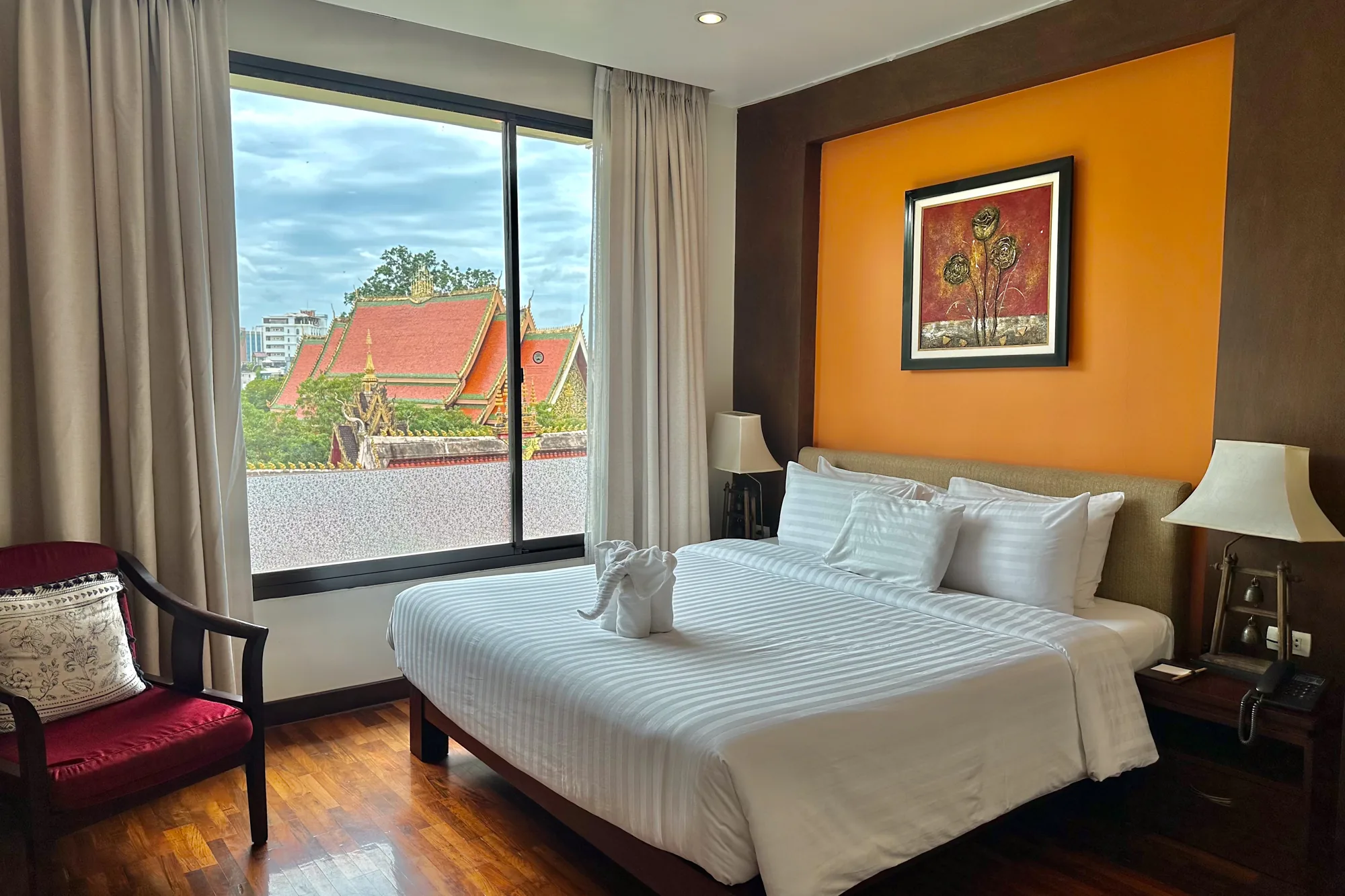 写真は、ラオスのビエンチャンにあるホテルの部屋を捉えています。部屋は明るく清潔感があり、窓からは寺院の屋根が見えます。 部屋の中心には、清潔な白いストライプのリネンがかけられたキングサイズのベッドがあります。ベッドの上には白い枕が重ねられ、タオルで作られた象の折り紙が置かれています。 ベッドのヘッドボードの後ろの壁は、オレンジ色と茶色のツートンカラーで塗られています。オレンジ色の部分には、額縁に入った絵が飾られています。ベッドサイドテーブルには、ランプと電話が置かれています。 窓からは、伝統的なラオス建築の寺院の屋根が見えます。窓のそばには、赤いクッションが置かれた木製の椅子があります。床は光沢のある木製で、部屋全体に温かみを与えています。全体的に、部屋は快適で魅力的な雰囲気です。