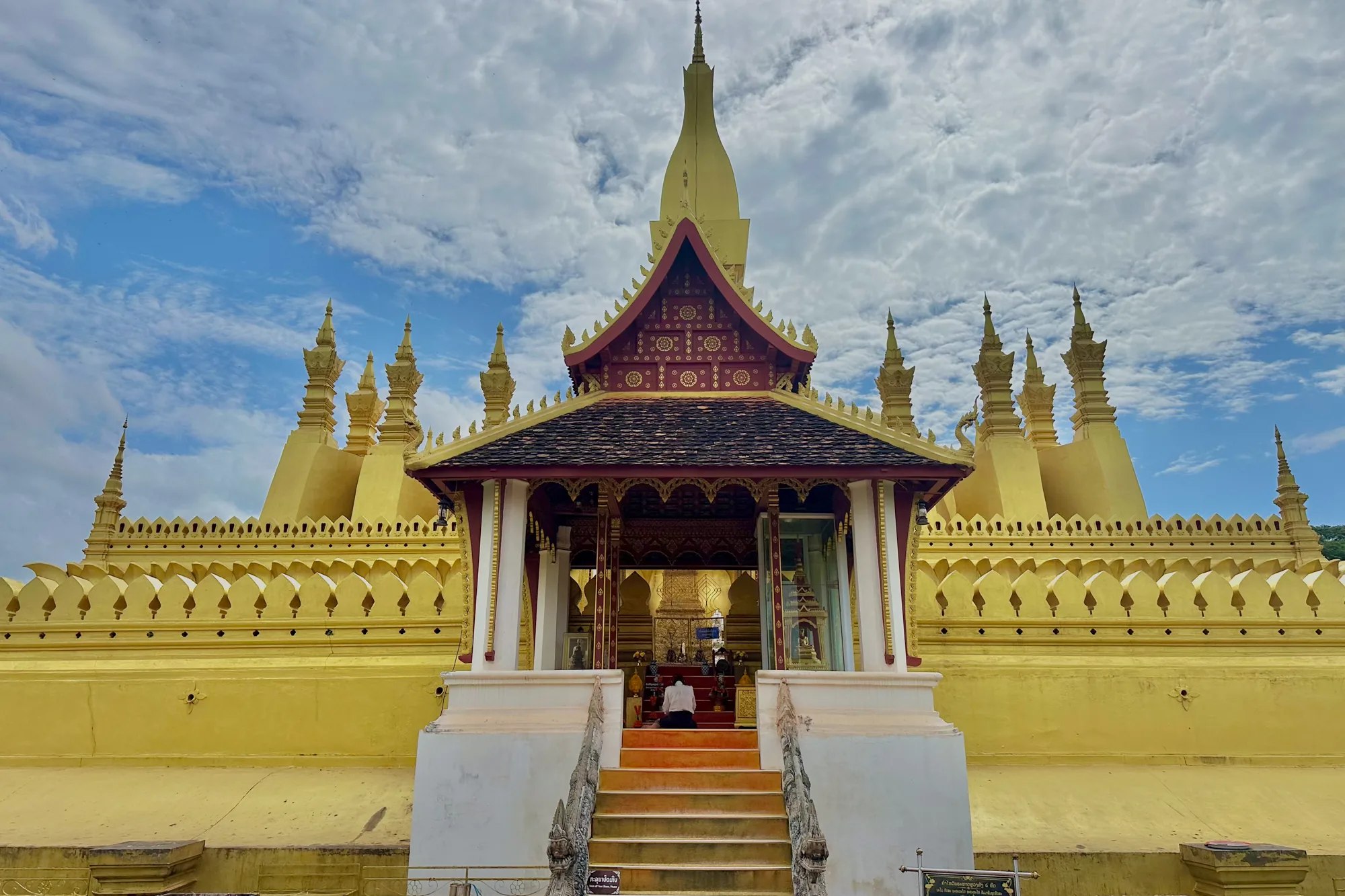 写真はラオスのヴィエンチャンで撮影されたもので、荘厳な黄金色の寺院が写っています。手前には赤い階段が続いており、その先に小さな堂があり、人がひざまずいて祈りを捧げている様子が確認できます。堂の屋根は黒い瓦で葺かれており、赤色の装飾が施されています。背景には巨大な黄金色の仏塔がそびえ立ち、その上部には特徴的な尖塔が空に向かって伸びています。空は青く、白い雲が点在し、穏やかな雰囲気を感じさせます。全体的に、写真からはラオスの文化的、宗教的な美しさと静寂が伝わってきます。