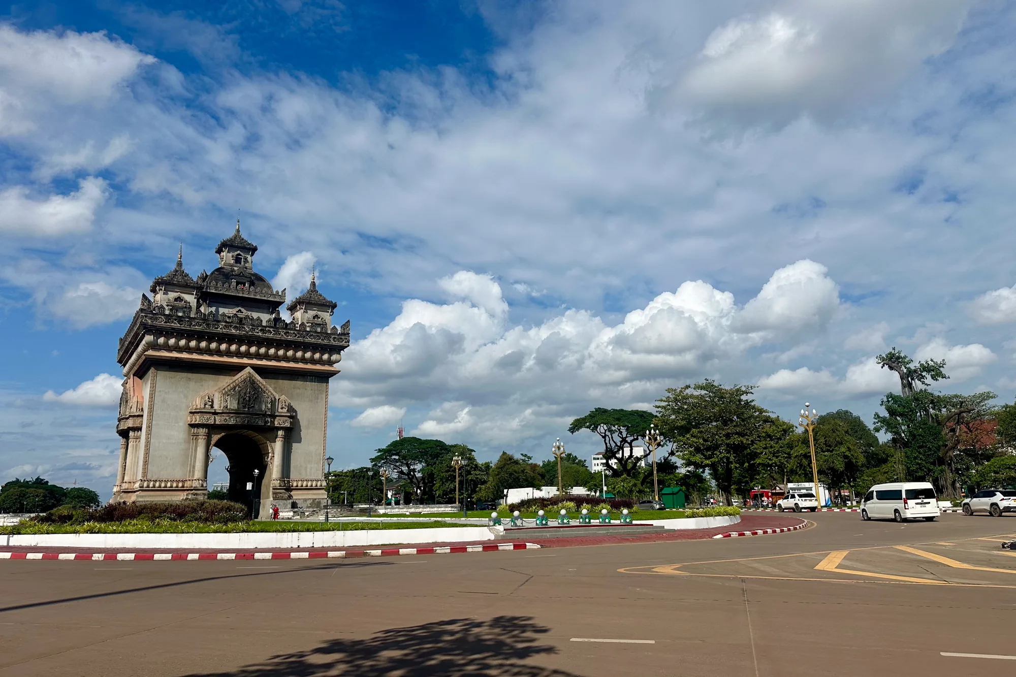 ラオスのビエンチャンで撮影された写真です。画面左側には、ラオスの象徴的な建造物であるパトゥーサイ（凱旋門）が堂々とそびえ立っています。石造りの重厚な造りで、細かな装飾が施されており、歴史を感じさせます。パトゥーサイの後ろには、青い空と白い雲が広がっており、そのコントラストが建物をより一層際立たせています。  パトゥーサイの周りはロータリーになっており、赤と白の縁石が道路を区切っています。画面右側には、緑豊かな木々が生い茂り、街灯が並んでいます。道路を走る車も写っており、活気のある様子が伺えます。全体的に、晴天の下でパトゥーサイが美しく際立つ、ラオスの風景を捉えた写真です。
