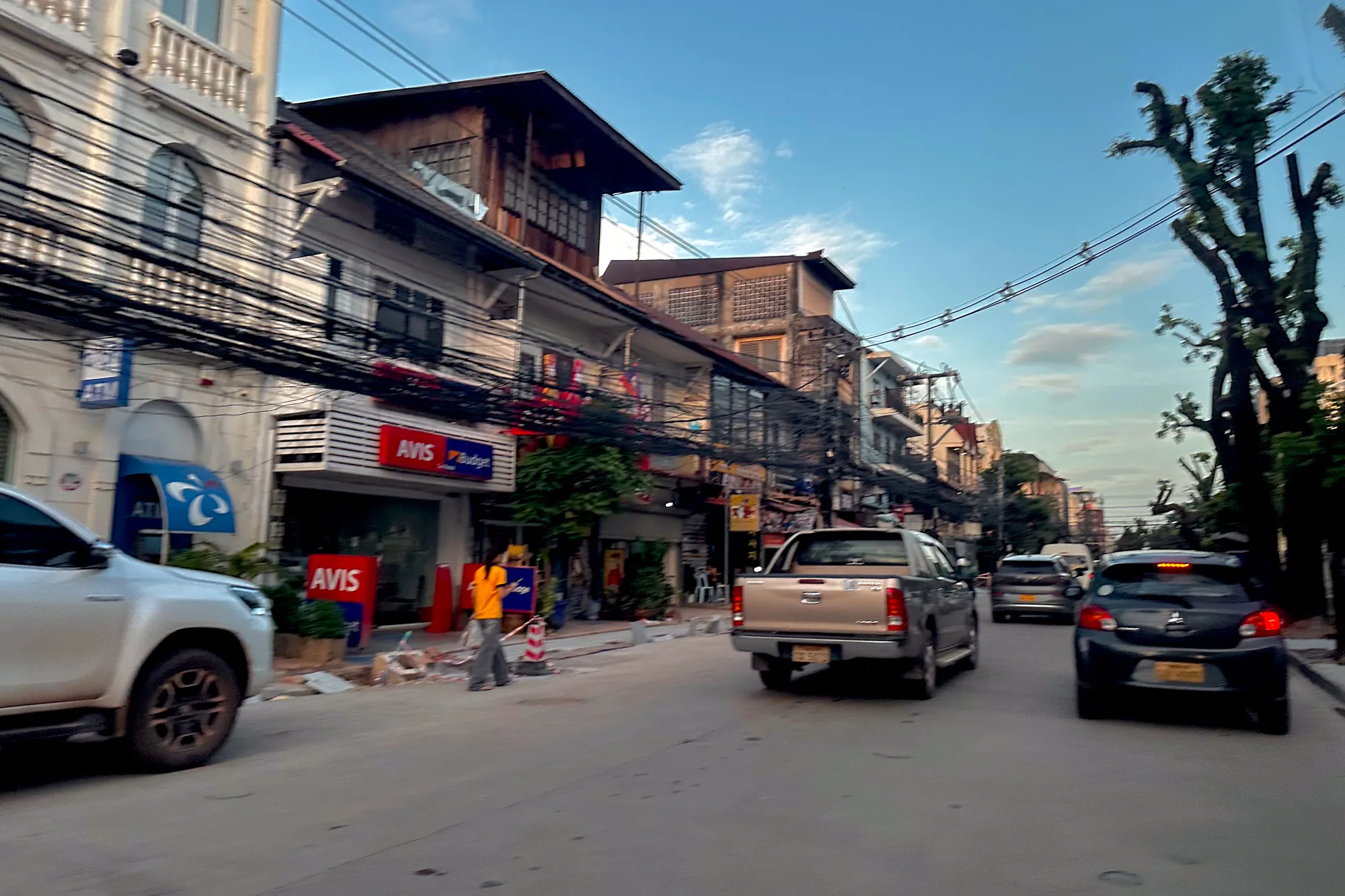 写真はラオスのビエンチャンで撮影されたものと思われます。画面中央やや左には、Avisのレンタカー店の看板が見えます。道路は舗装されており、画面右方向へ向かって伸びています。路上には数台の自動車が走行しており、手前からシルバーのSUV、シルバーのピックアップトラック、黒いハッチバック車などが確認できます。道路脇では作業員がオレンジ色のシャツを着て作業をしているようです。電線が複雑に張り巡らされており、東南アジアの街並み特有の風景を作り出しています。建物は古く、植民地時代の建築様式を残しているものも見受けられます。空は晴れており、薄い雲が浮かんでいます。全体的に、穏やかな午後の光が差し込むビエンチャンの日常風景を切り取った写真と言えるでしょう。