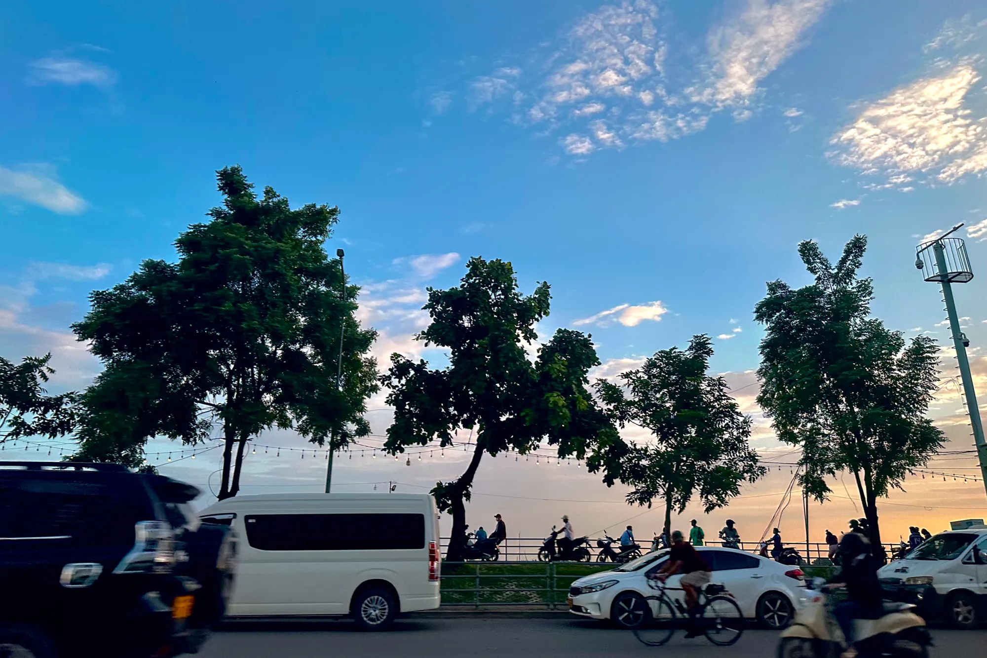 ラオスのビエンチャンで撮影された写真です。夕暮れ時の空が美しく、青とオレンジのグラデーションが印象的です。空には薄い雲が広がっています。写真には、道路沿いに並ぶ木々、行き交う車やバイク、そして夕涼みを楽しむ人々のシルエットが写っています。路上には黒い車と白いバン、白い車が写っており、自転車に乗った人も写っています。遠くの川沿いには人が並んでいて、夕暮れを楽しんでいるようです。上空には電飾が連なっており、夕暮れの雰囲気をさらに盛り上げています。全体的に落ち着いた雰囲気で、ビエンチャンののどかな日常風景が伝わってきます。