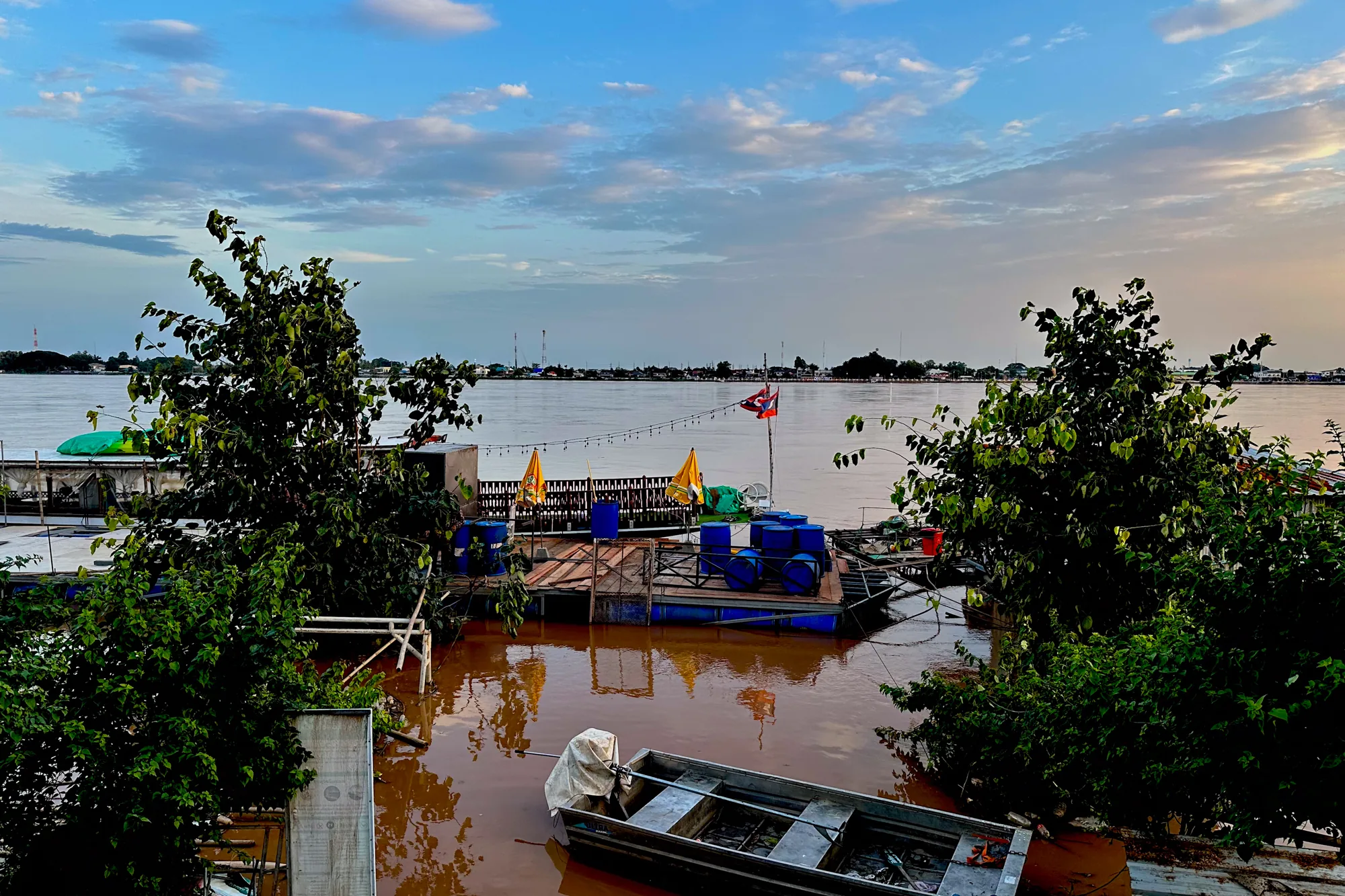ラオスのビエンチャンで撮影された写真です。夕暮れ時のメコン川が写っています。空は青色と薄いオレンジ色に染まり、雲が浮かんでいます。手前には緑豊かな木々があり、その奥に茶色く濁ったメコン川が広がっています。川には、浮かんでいるプラットフォームやボートが見えます。プラットフォームには青い樽や黄色のパラソル、ラオスの国旗が立っています。ボートは水に浸かりかけており、船体の一部が白いシートで覆われています。対岸には建物や電波塔が立ち並び、そのシルエットが夕焼け空に浮かび上がっています。全体的に、静かで穏やかな雰囲気の写真です。