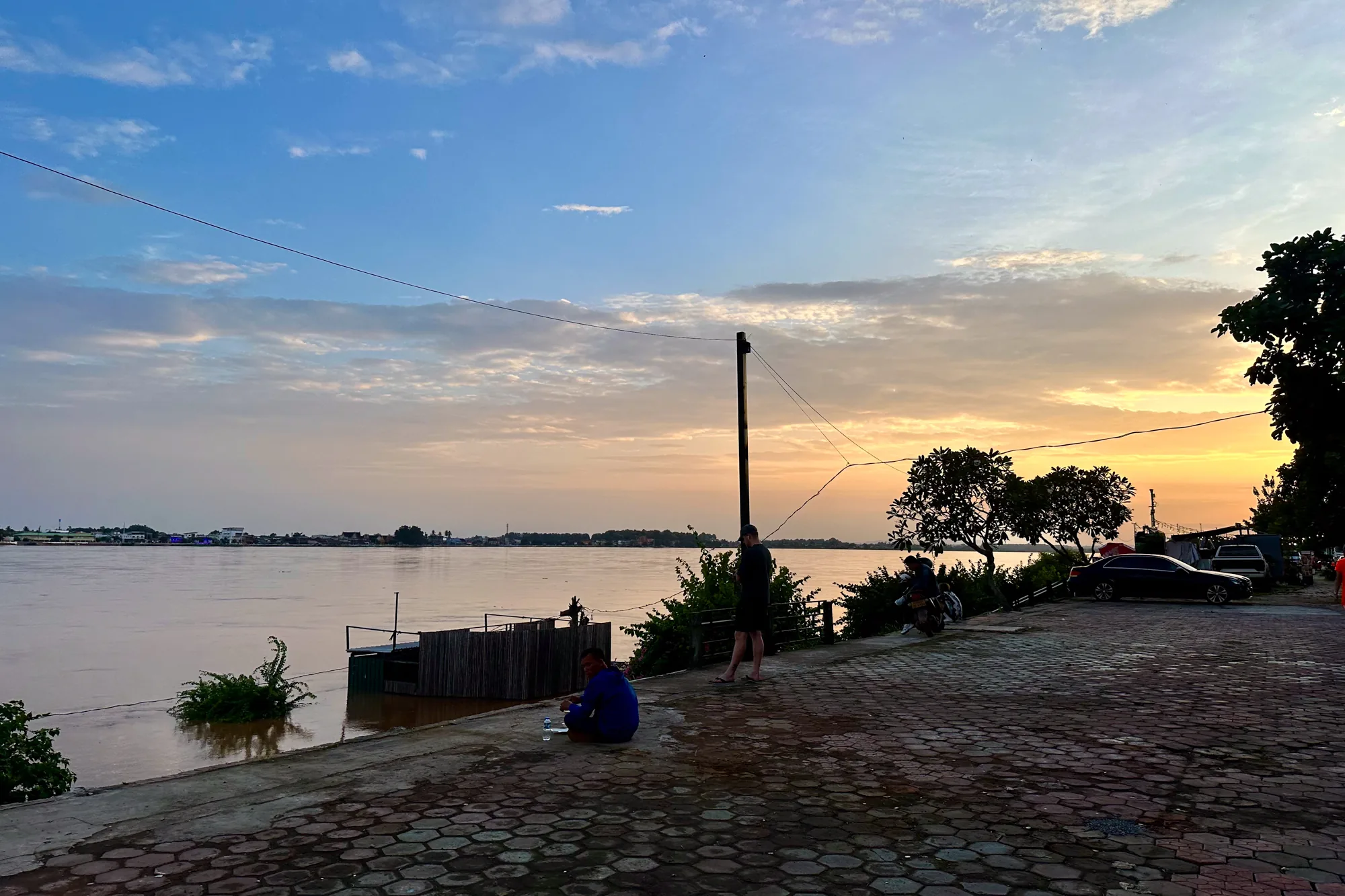 ラオスのビエンチャンで撮影された、夕暮れ時のメコン川の風景写真です。空は、青色から夕日のオレンジ色へとグラデーションがかかっており、美しい雲が広がっています。川の水面は穏やかで、夕焼けの色を反射して茶色っぽく見えます。対岸には建物が並んでいるのが見えます。手前には石畳の道があり、人々がくつろいでいます。ブルーのシャツを着た男性が座って何か飲んでおり、立っている人やバイクに座っている人もいます。夕方の穏やかな時間が流れている様子が伝わってきます。電線が空を横切っており、生活感を感じさせます。道路脇には木々が茂り、車も数台駐車しており、日常の風景を切り取った写真と言えるでしょう。