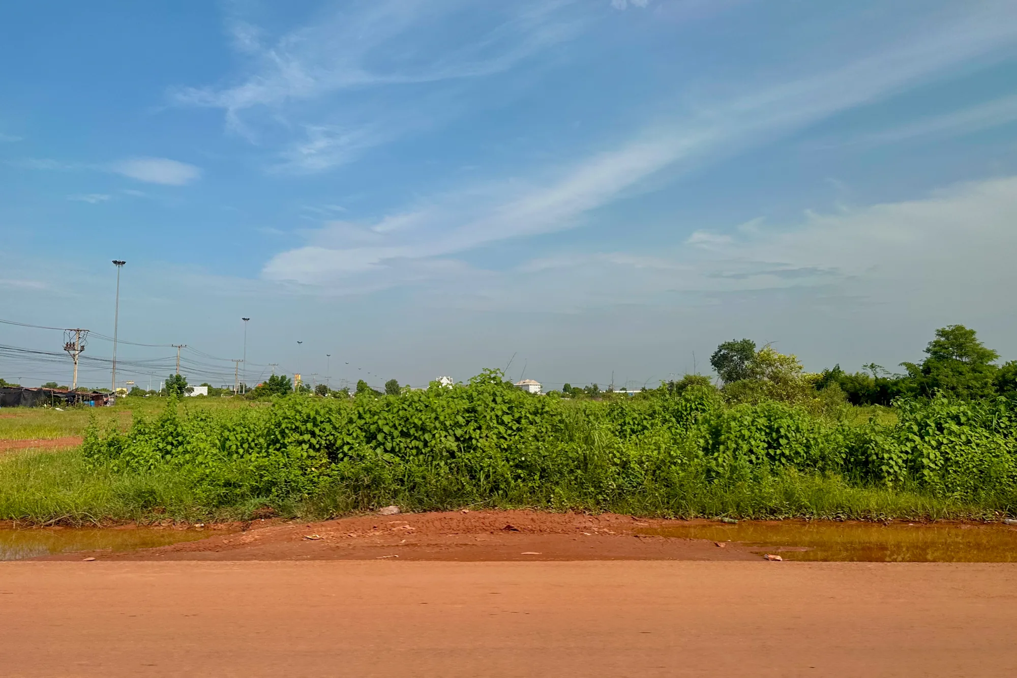 写真はラオスのビエンチャンで撮影されたもので、広々とした青空と緑豊かな風景が印象的です。画面手前には赤茶色の土が露出した道があり、その向こうには生い茂った草木が広がっています。草木は緑色の葉を茂らせ、生命力にあふれています。奥には電柱が並び、電線が空を横切っているのが見えます。遠景には白い建物らしきものも確認できます。空は青く、白い雲が点在しており、穏やかな天候を物語っています。全体的に、のどかで静かな雰囲気が感じられます。東南アジア特有の赤土と、豊かな緑が混ざり合った風景が、ビエンチャンの素朴な魅力を伝えています。