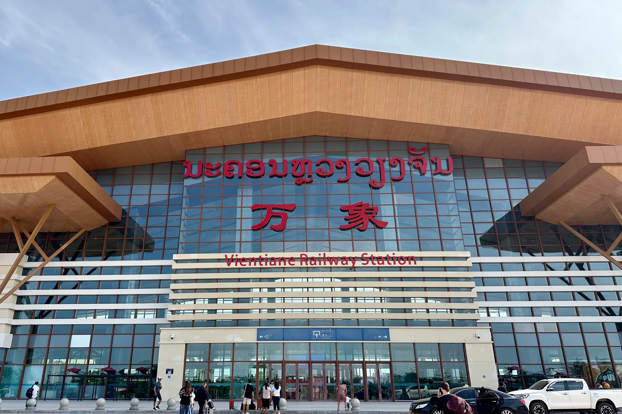 これはラオスのヴィエンチャン駅の写真です。駅舎はモダンなデザインで、ガラス張りの外観と木製の軒が特徴的です。建物の上部にはラオス語、中国語、英語で駅名が書かれています。駅の前には人々が歩いており、タクシーや自家用車が停車しています。空は晴れており、日中の撮影であることがわかります。全体的に、新しく近代的な鉄道駅の印象を与える写真です。