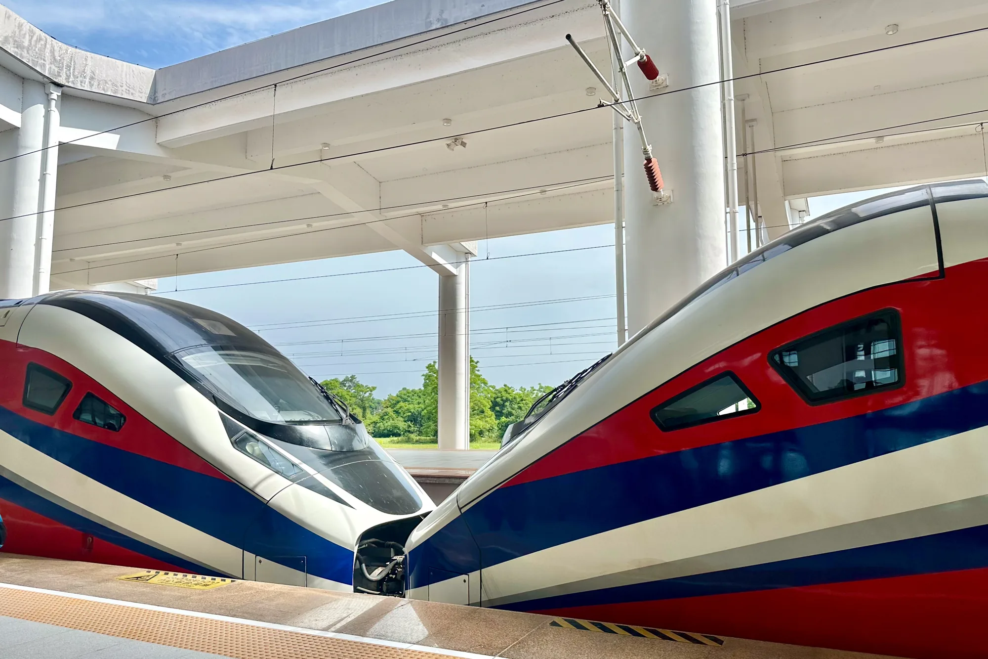 ラオスの首都ビエンチャンで撮影された写真です。駅のホームに停車している高速鉄道の車両が2つ写っています。車両は赤、白、青のストライプで彩られており、流線型のデザインが特徴的です。車両の先頭部分は互いに向き合うように配置されています。背景には駅の構造物や架線柱、そして緑豊かな木々が見えます。明るい日差しが降り注いでおり、近代的な鉄道インフラとラオスの自然が組み合わさった様子が印象的です。