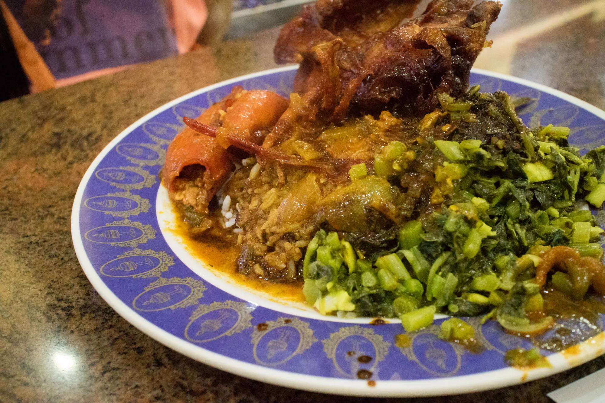 写真には、マレーシアのクアラルンプールで撮影されたと思われる料理が盛られた皿が写っています。皿は濃い青色で、金色の装飾が施されています。料理はご飯の上に様々な種類の具材が載せられており、いくつかのおかずが添えられています。左側には赤いパプリカのようなものに詰め物をした料理、中央には肉や野菜を煮込んだような料理、右側には細かく刻んだ緑色の野菜炒めのようなものが確認できます。また、奥には揚げた肉のようなものも見えます。全体的に、マレーシアの伝統的な料理を盛り合わせたプレートのようです。照明の関係か、料理の色味が少し暗く、食欲をそそるような鮮やかさには欠けるかもしれません。