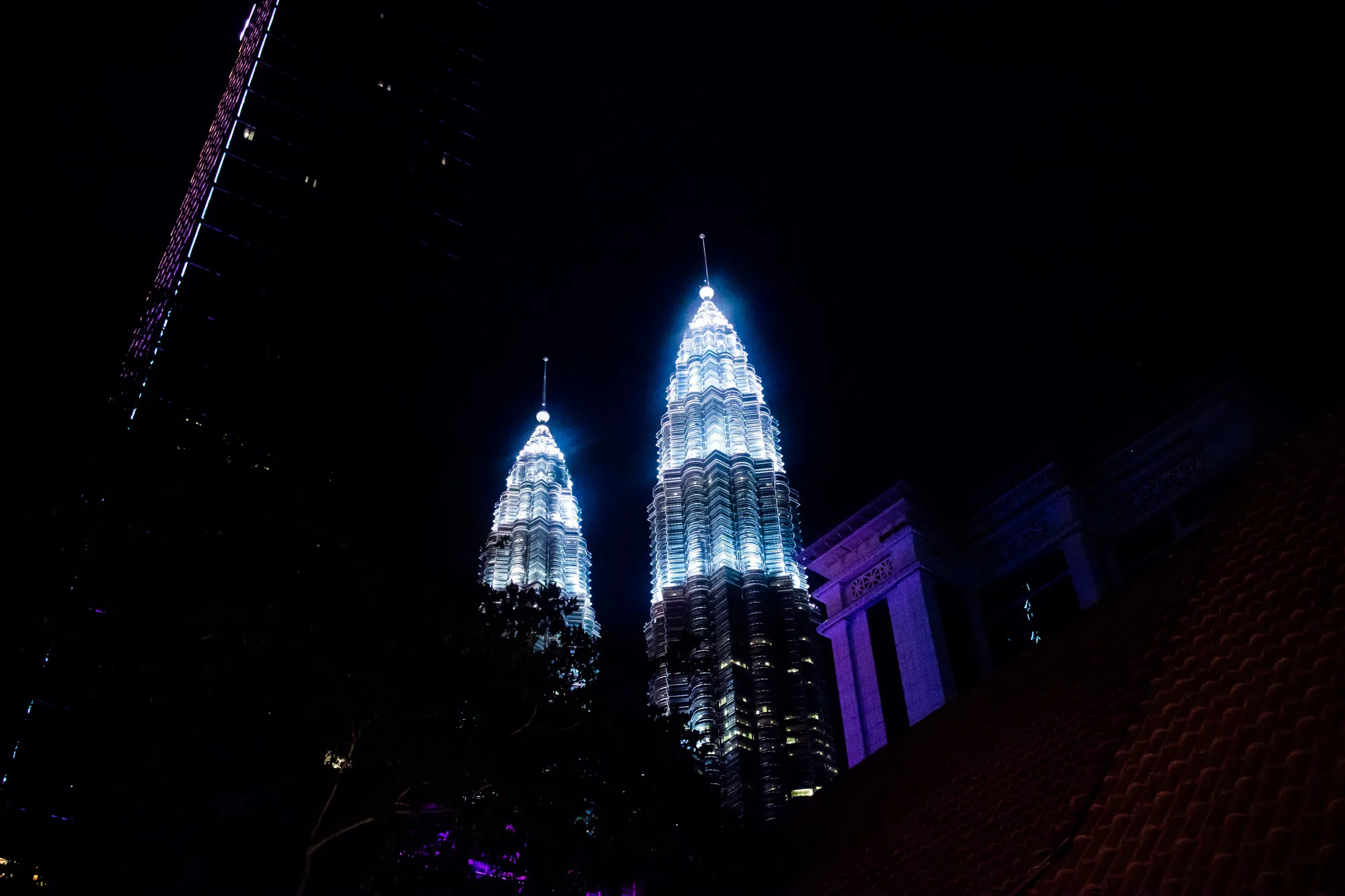 写真はマレーシアのクアラルンプールで撮影された、夜のペトロナスツインタワーを捉えたものです。周囲は暗闇に包まれており、その中でツインタワーが白と青のライトアップによって際立っています。タワーの頂上部分は明るく輝き、細部まで精巧な建築構造が浮かび上がっています。  画面左側には、一部がフレームインした別の高層ビルが見えます。また、手前には木々のシルエットが重なり、夜景に奥行きを加えています。画面右側には、紫色のライトアップが施された建物の一部と、赤茶色の屋根瓦が写っており、伝統的な要素が夜景に彩りを添えています。全体的に、都会の夜の美しさと静けさを感じさせる写真です。