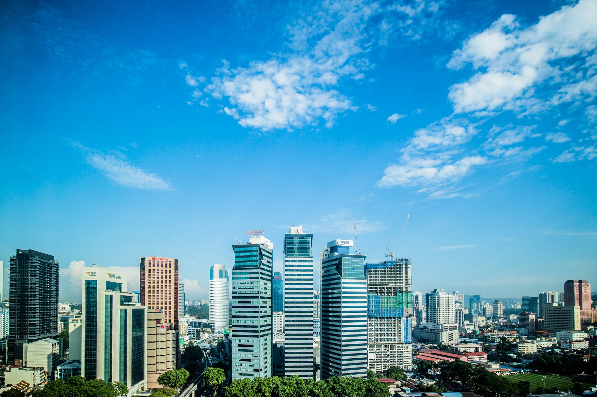 写真は、マレーシアのクアラルンプールで撮影された都市の風景を捉えています。空は鮮やかな青色で、白い雲が点在しており、爽やかな印象を与えます。手前には緑豊かな木々が茂り、その奥には様々な高さのビルが立ち並ぶ様子が広がっています。モダンな高層ビルから、特徴的なデザインのビル、建設中のビルまで、多様な建築物が確認できます。街並みは整然としており、近代的な都市の活気を感じさせます。写真全体は明るく、晴れやかな雰囲気で、クアラルンプールの都市景観の美しさを際立たせています。