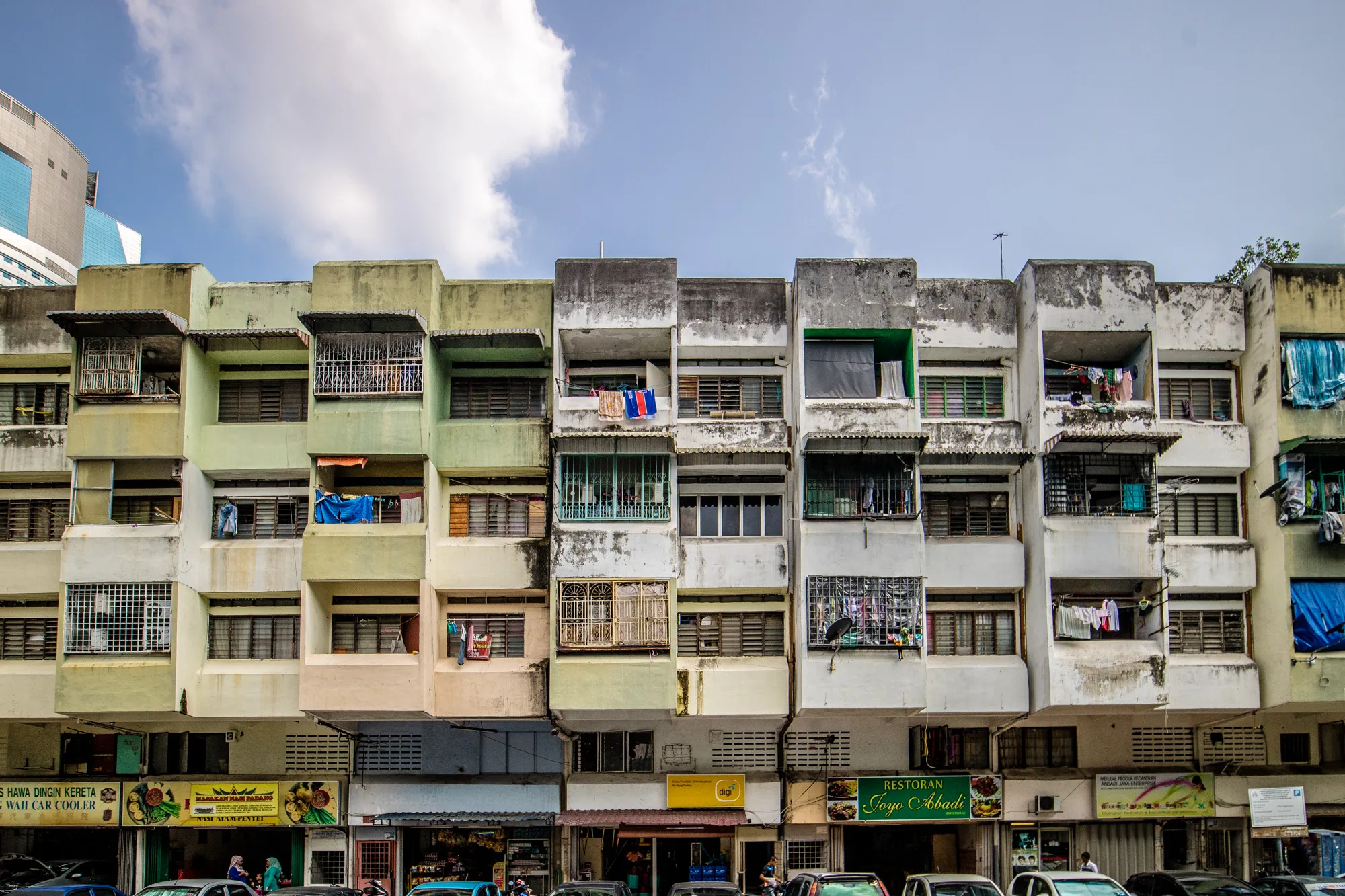 写真はマレーシアのクアラルンプールで撮影されたと思われる建物群を捉えています。建物は複数階建てで、外観はやや古びた印象です。様々な色合いの壁面が連なり、窓には鉄格子が取り付けられているものもあります。ベランダには洗濯物が干されており、生活感があふれています。建物の下には店舗が並び、看板にはローカルなレストランの名前（Restoran Joyo Abbadiなど）が見られます。路上には車が駐車しており、賑わいを感じさせます。空は一部雲がかかっていますが、全体的に明るい日中の写真です。