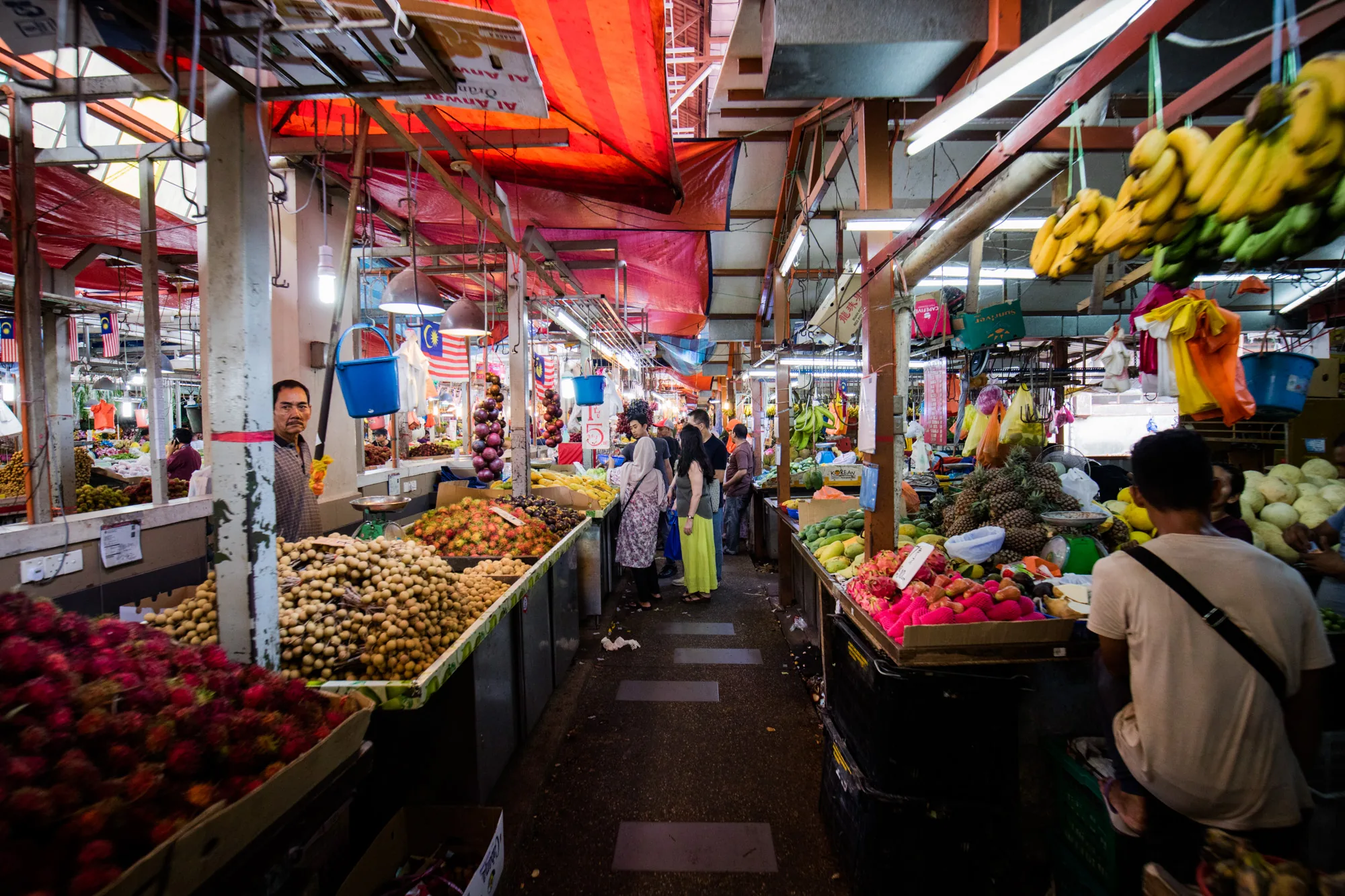 写真はマレーシアのクアラルンプールにある市場の様子を捉えたものです。通路の両側には様々な果物や野菜を並べた屋台が連なり、多くの人々が買い物に訪れています。手前には赤色のランブータンや茶色のロンガンが山積みにされており、その奥には玉ねぎやトウモロコシ、他の種類の果物が並んでいます。通路には買い物客が行き交い、活気に満ちた雰囲気です。天井には赤いシートやマレーシアの国旗が飾られており、蛍光灯が市場全体を明るく照らしています。通路奥にはバナナが吊るされており、トロピカルな雰囲気を醸し出しています。カメラに向かってこちらを見ている店員の表情からは、市場の日常が感じられます。全体的に、色彩豊かでエネルギッシュな市場の様子が伝わってくる写真です。