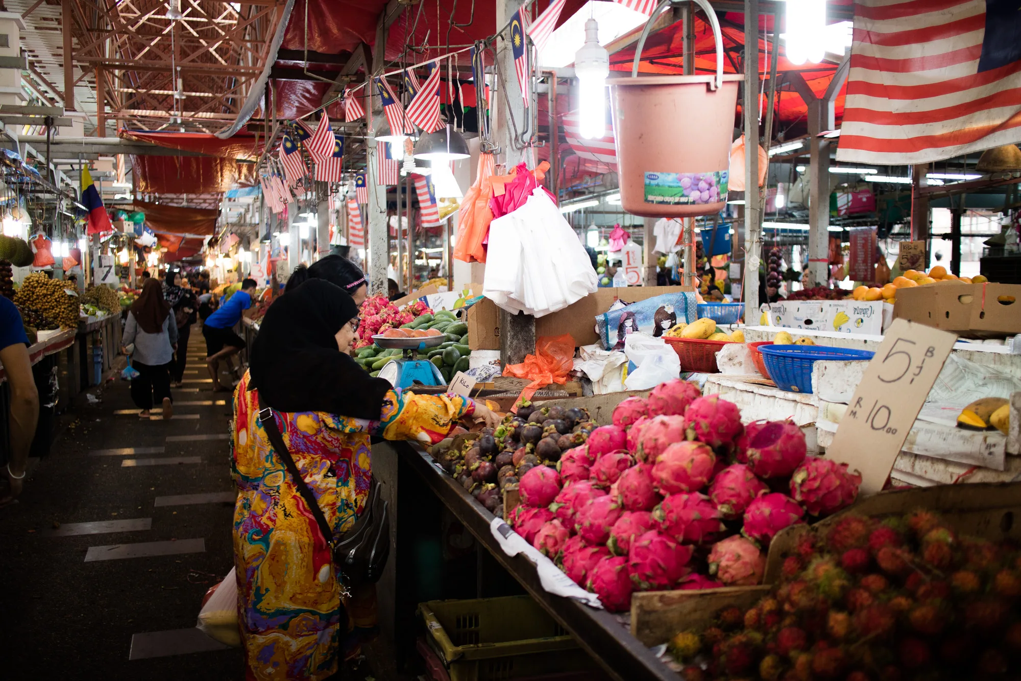 この写真はマレーシアのクアラルンプールで撮影された市場の様子を捉えています。屋内市場の活気ある雰囲気が伝わってきます。天井は金属製の骨組みで覆われており、赤色の覆いが一部にかけられています。マレーシアの国旗が天井から吊り下げられており、ローカルな雰囲気を強調しています。  写真には、ヒジャブを被り、鮮やかな色彩の服を着た女性が写っています。彼女は果物の品定めをしているようです。市場の屋台には、ドラゴンフルーツ、マンゴスチン、ランブータンなど、様々な種類の果物が山積みにされています。価格を示す手書きの看板が見えます。  背景には、買い物客が行き交う様子が見えます。全体的に、ローカルな市場の活気と賑わいが感じられる写真です。