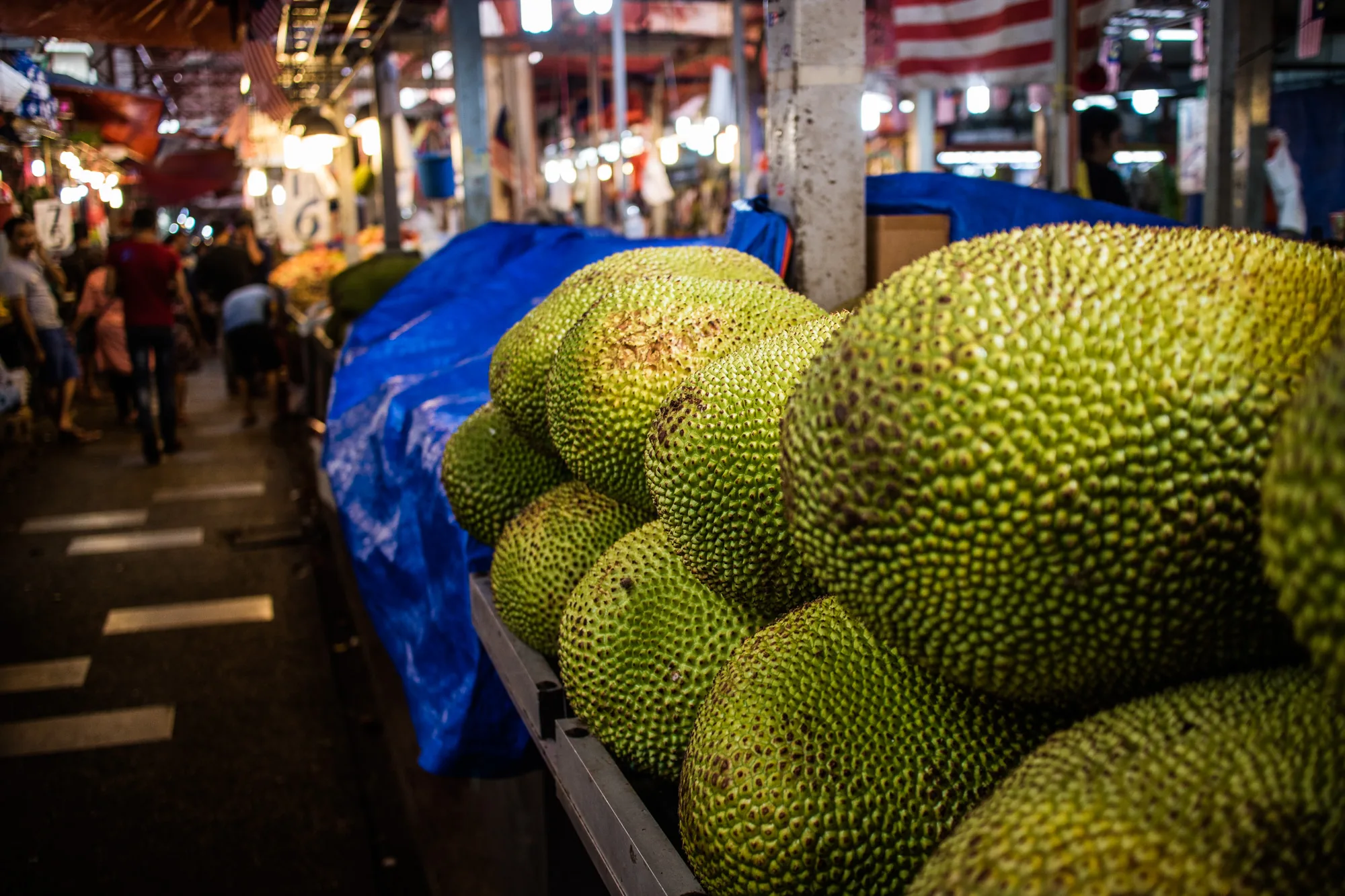 写真はマレーシアのクアラルンプールで撮影された、活気ある市場の様子を捉えています。前景には、青いシートの上に山積みにされた大きな緑色のジャックフルーツが並んでいます。ジャックフルーツの表面はイボイボしており、独特の質感が際立っています。背景には、多くの人々が行き交う様子が写っており、市場の賑わいが感じられます。店舗には、マレーシアの国旗である「ジャラル・グミルアン」が飾られており、現地の雰囲気を醸し出しています。頭上には電球が灯り、市場全体を明るく照らしています。写真全体は、東南アジアの市場特有の熱気と活力を伝えています。