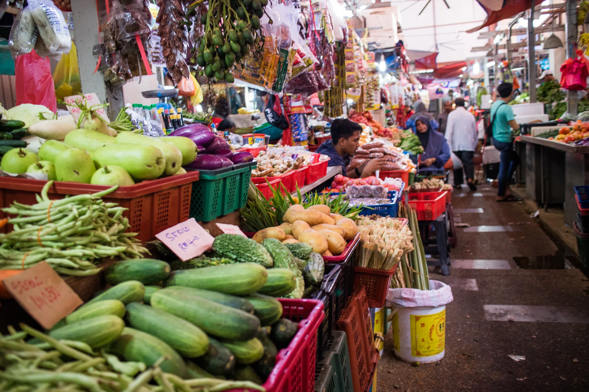 この写真はマレーシアのクアラルンプールにある市場の様子を捉えています。画面手前には、様々な種類の野菜が所狭しと並べられています。キュウリ、インゲン、冬瓜、ナスなど、色とりどりの野菜がプラスチック製のカゴに盛られています。それぞれの野菜には手書きの価格表示札が添えられています。奥には、果物や乾物などを売る店が連なり、多くのお客さんで賑わっています。店員の男性が商品を並べたり、お客さんとやり取りしている様子が見えます。通路はやや薄暗く、湿気を含んだ空気感が伝わってきます。天井には屋根があり、ところどころに明かりが灯っています。アジアの市場特有の活気と熱気が感じられる写真です。