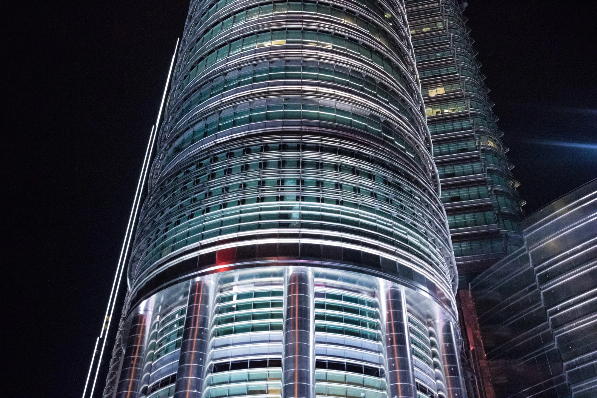 写真は、マレーシアのクアラルンプールにあるペトロナスツインタワーを夜間に撮影したものです。ローアングルから見上げており、そびえ立つタワーの威容が強調されています。暗闇を背景に、タワーのガラス張りの壁面と金属製の構造がライトアップされ、未来的で洗練された雰囲気を醸し出しています。タワーの曲線的なデザインと、水平方向に走るラインが、光と影のコントラストを生み出し、独特の美しさを際立たせています。一部の窓からは室内の光が漏れており、タワー内部の活動をほのかに感じさせます。全体的に、都市の夜景を象徴する印象的な一枚です。