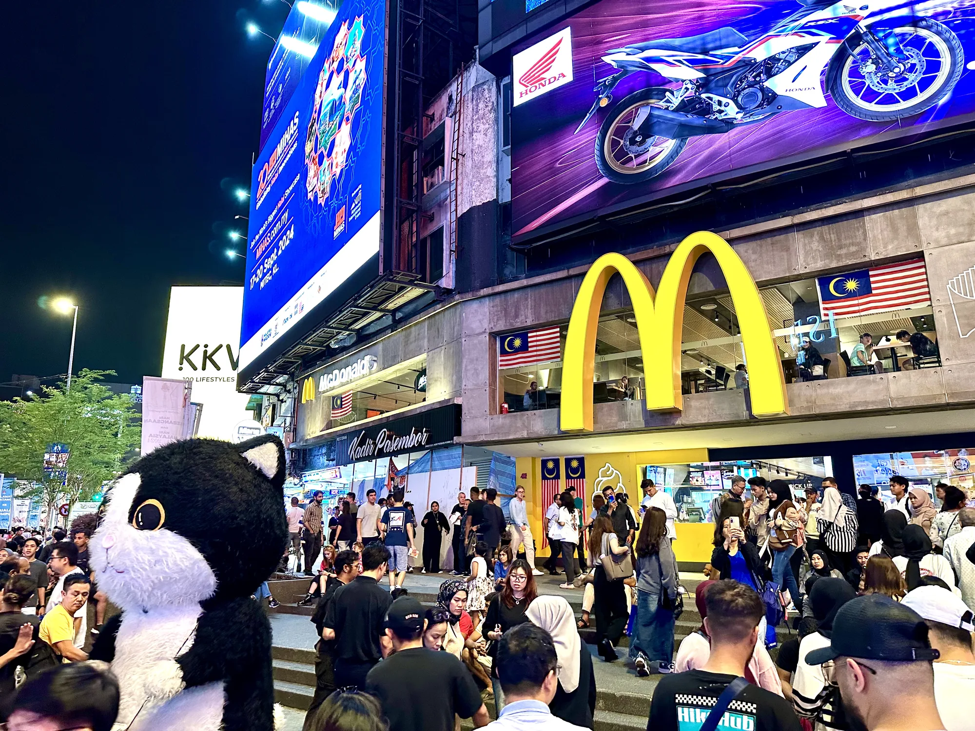 写真はマレーシアのクアラルンプールで撮影された夜の風景です。大勢の人々が行き交う賑やかな通りが写っています。手前には黒と白の猫のキャラクターの着ぐるみが見えます。背景にはマクドナルドの店舗があり、マレーシアの国旗が掲げられています。店舗の上には大きなゴールデンアーチのロゴが輝いています。  さらにその上には、ホンダのバイクの広告が大きく表示されています。左側には「KKV」と書かれた看板や、「MIHAS.com.my」と書かれた大きな広告が立っています。これらの広告は夜の街を明るく照らし、活気ある雰囲気を醸し出しています。通りには様々な服装をした人々がおり、観光客や地元の人々が混ざり合っている様子が伺えます。全体的に、クアラルンプールの賑やかでエネルギッシュな夜の雰囲気が伝わってくる写真です。