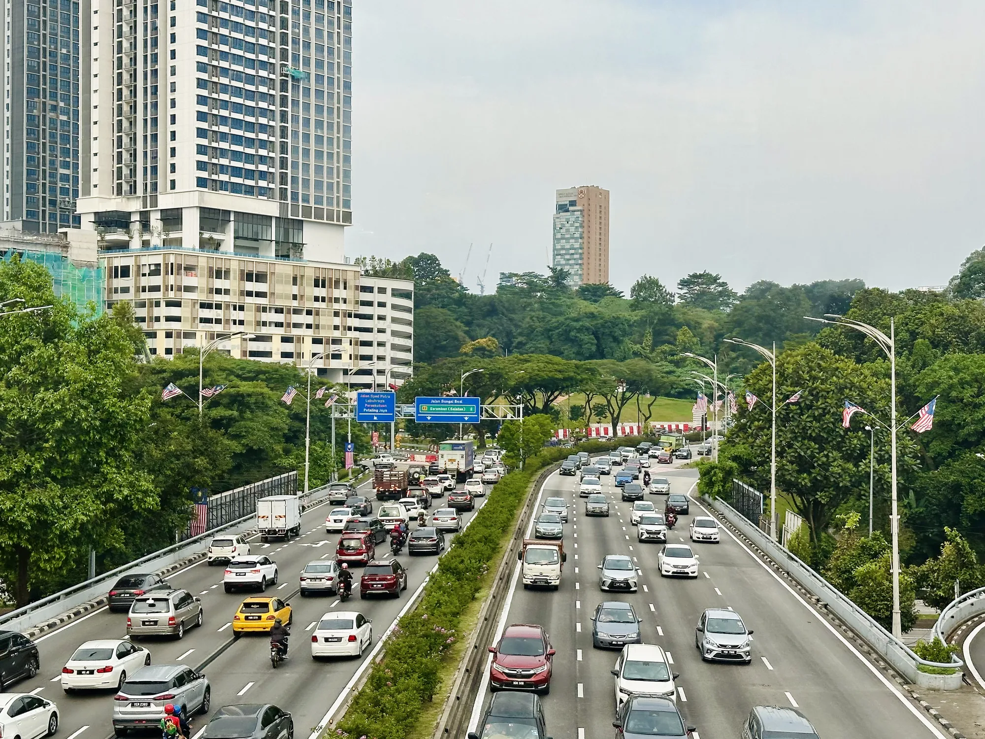 写真は、マレーシアのクアラルンプールで撮影された、交通量の多い高速道路の様子を捉えています。 高層ビルが背景に見え、緑豊かな木々が道路の両側に生い茂っています。空は曇り気味で、全体的にやや霞んだ雰囲気です。道路は複数の車線に分かれており、自動車、トラック、バイクなど、さまざまな種類の車両がひしめき合っています。交通は比較的混雑しており、速度は遅いようです。マレーシアの国旗が道路沿いに掲げられており、場所を特定するのに役立ちます。全体的に、都市の喧騒と交通渋滞をリアルに伝える写真です。