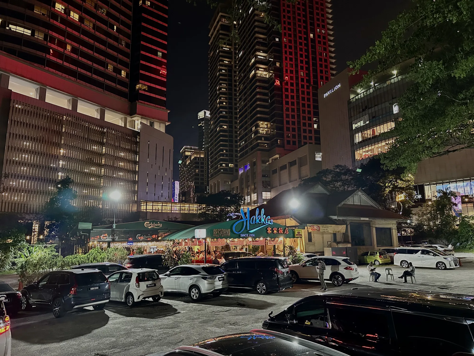 この写真は、マレーシアのクアラルンプールで撮影された夜景です。手前には駐車場があり、さまざまな車種の車が多数駐車されています。駐車場には人影もいくつか見えます。奥にはレストランらしき建物があり、「Hakka」という大きな青い看板が印象的です。「SINCE 1956」と書かれていることから、老舗のレストランであることがわかります。レストランの屋根にはマレーシア国旗が飾られており、多くの人が食事を楽しんでいる様子がうかがえます。さらに奥には高層ビルが立ち並び、建物の一部が赤くライトアップされています。写真全体としては、活気あるクアラルンプールの夜の街並みを捉えた一枚と言えるでしょう。
