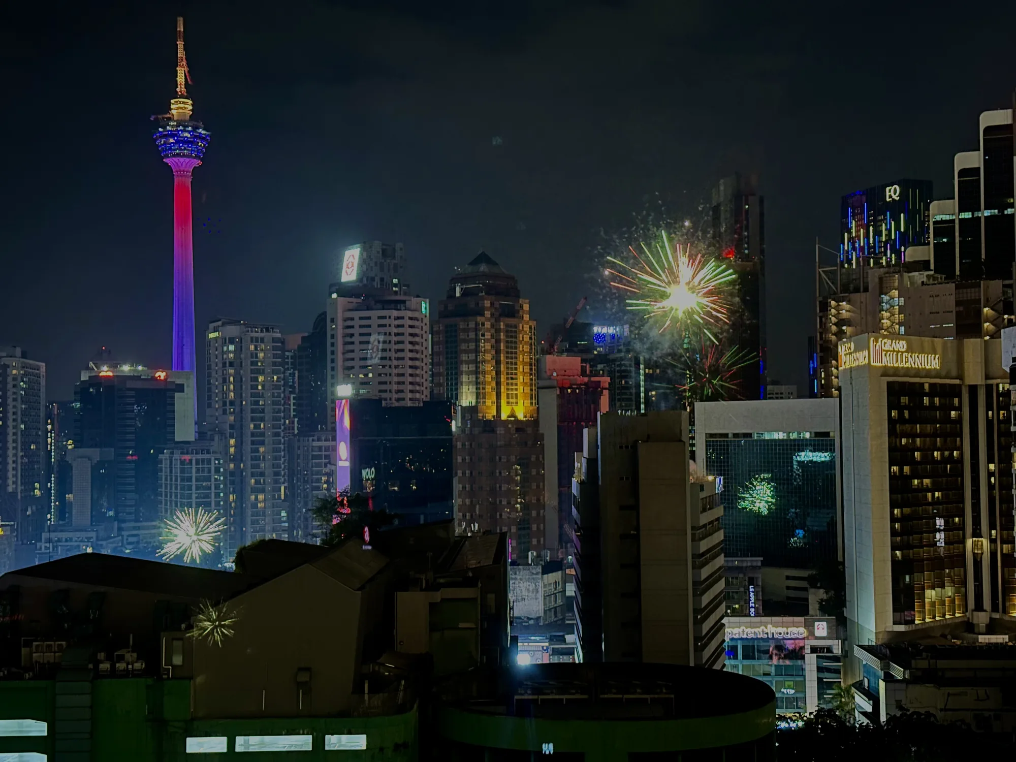 写真はマレーシアのクアラルンプールで撮影された夜景です。高層ビル群が立ち並び、夜空には花火が打ち上がっています。手前に見えるのは、緑色の屋根の建物の一部です。  画面左側には、赤と青のライトアップが施されたクアラルンプールタワーがそびえ立っています。他の高層ビルもライトアップされており、それぞれの建物が異なる色やデザインで輝いています。例えば、「GRAND MILLENNIUM」と書かれたホテルや、「EQ」のロゴが光るビルが見えます。  空には花火がいくつか打ち上げられており、特に中心部に見える大きな花火は緑や赤の光を放ち、夜景をさらに華やかに彩っています。全体的に、都会の賑やかさと祝祭感を感じさせる写真です。
