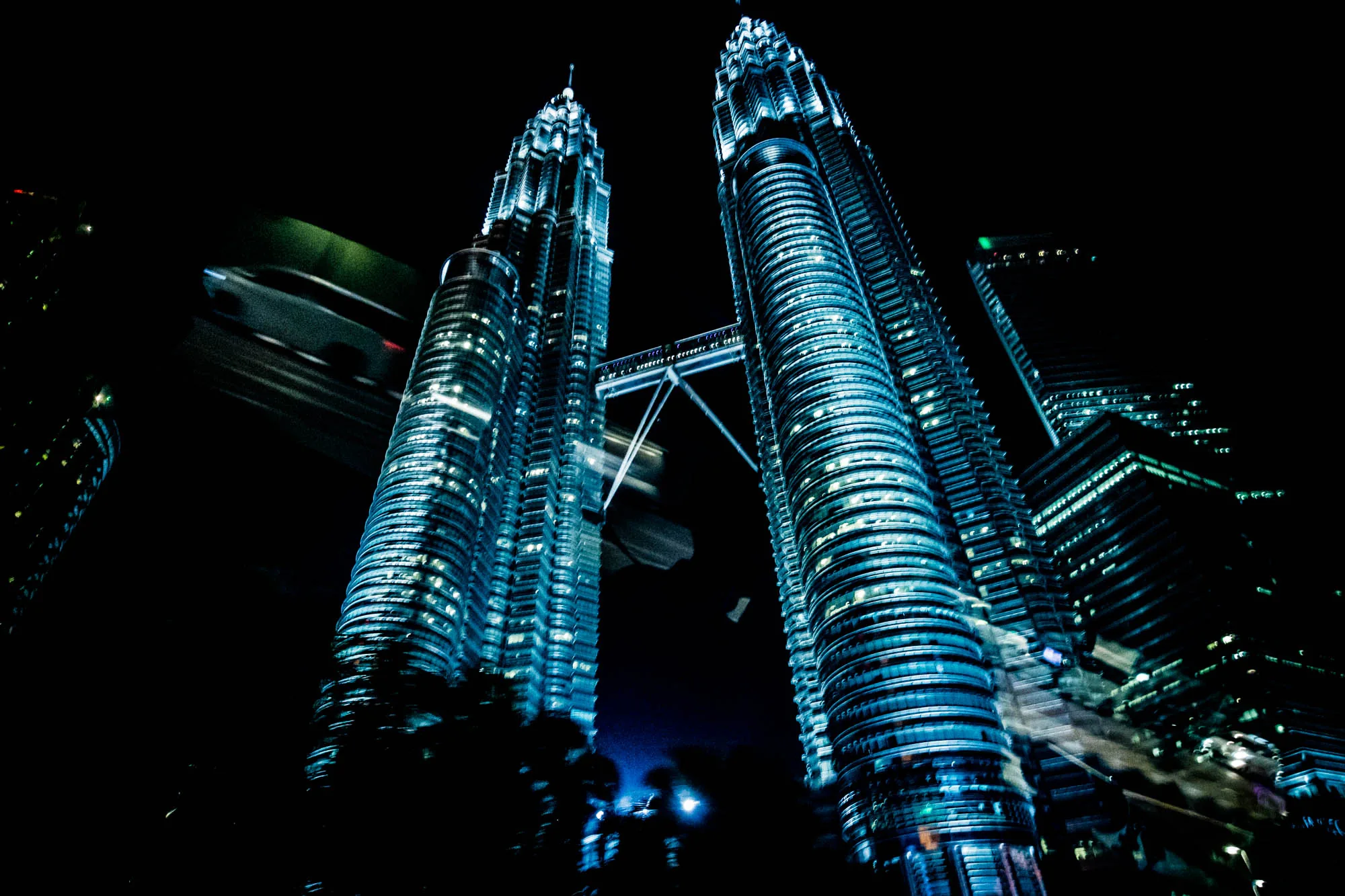マレーシアのクアラルンプールで撮影された夜景写真です。画面中央には、青白い光を放つペトロナスツインタワーがそびえ立っています。二つのタワーはスカイブリッジで結ばれており、その存在感が際立っています。手前には樹木のシルエットが写り、夜の闇の中で浮かび上がっています。左上には、高速で移動する車両の光跡がぼやけて写り込み、都市の活気を伝えています。全体的に青みがかった色調で、未来的な雰囲気が強調されています。他の高層ビルも写り込んでおり、都会の夜を象徴するような光景です。
