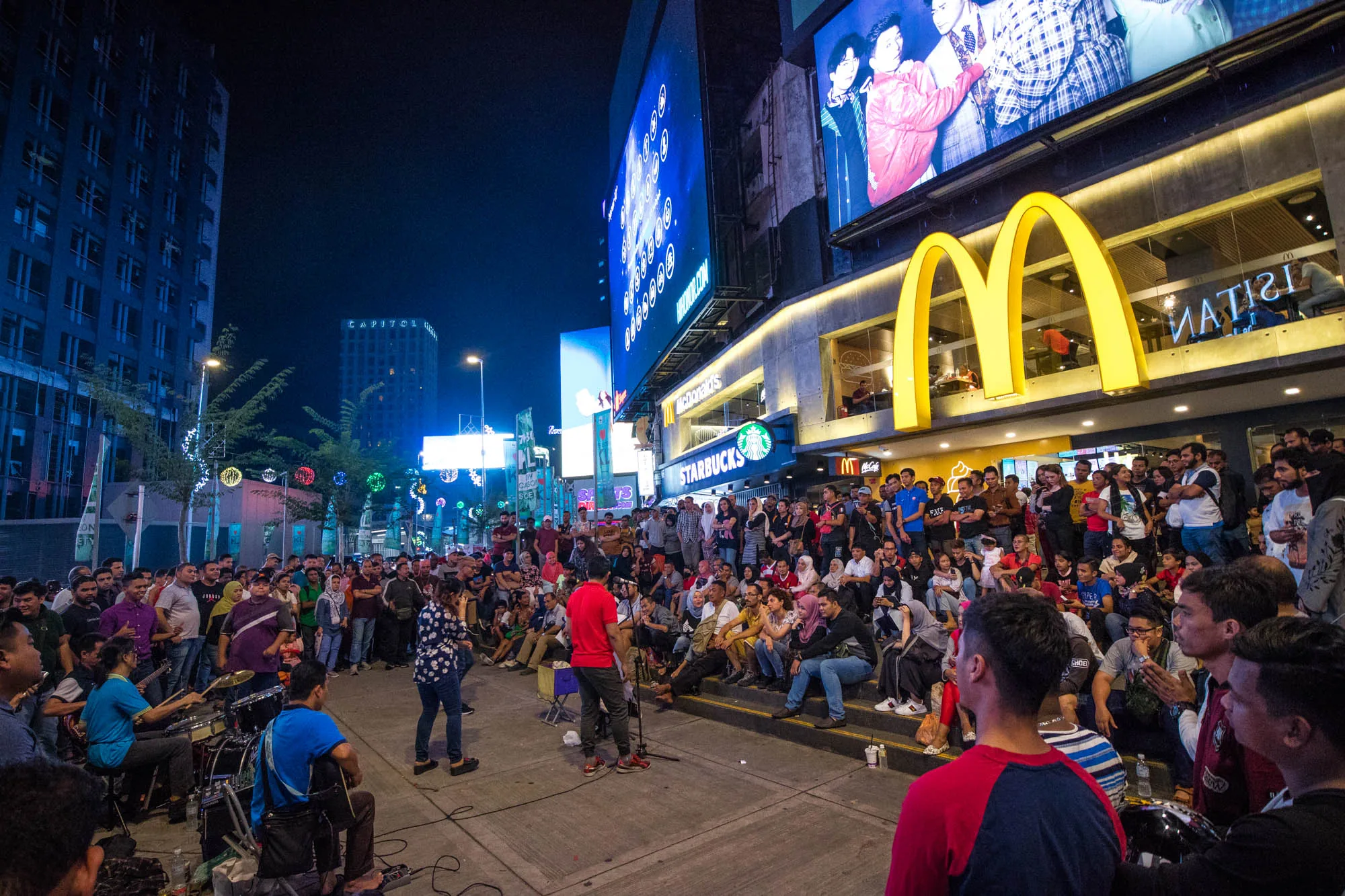 この写真はマレーシアのクアラルンプールで撮影された夜景です。大勢の人々が集まり、賑わっている様子が写っています。画面中央には、人々が座って鑑賞している階段状のスペースがあり、その前には路上ミュージシャンが演奏しています。バンドはギター、ドラム、キーボードで構成され、音楽を奏でています。人々は演奏に耳を傾け、リラックスした雰囲気です。  背景には、マクドナルドとスターバックスの看板が明るく光り、ネオンサインが周囲を照らしています。ビルの壁面には大きな広告が掲示されており、クアラルンプールの都会的な雰囲気を醸し出しています。さらに奥には、高層ビル群が立ち並び、「CAPITOL」と書かれた建物も見えます。街路樹にはイルミネーションが施され、夜の街に彩りを添えています。全体的に、賑やかで活気のあるクアラルンプールの夜の様子が伝わってくる写真です。