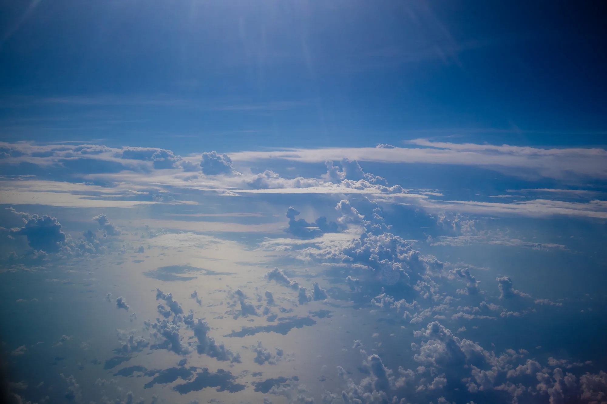 写真は、マレーシアのクアラルンプール上空で撮影されたと思われる、空の景色を捉えています。画面の上部は濃い青色の空が広がり、太陽光がかすかに差し込んでいます。中層から下層にかけては、大小さまざまな白い雲が層をなし、雲海を形成しています。雲の形は多様で、もくもくと盛り上がったものや、薄くたなびくものなどが見られます。全体的に青と白の色調が美しく、空の広大さと雲の繊細さを感じさせる一枚です。