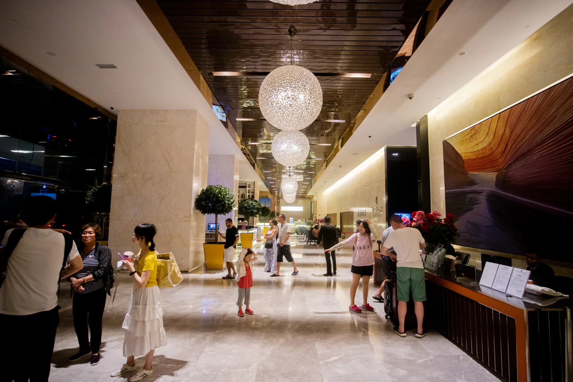 これはマレーシアのクアラルンプールで撮影された写真です。写真には、高級ホテルのロビーが写っています。ロビーは広々としており、大理石の床が磨き上げられています。天井には、多数の球体のシャンデリアが並んでおり、豪華な雰囲気を醸し出しています。ロビーには、チェックインカウンターがあり、数人の宿泊客が手続きをしています。その他にも、談笑する人々、通り過ぎる人々など、様々な人が写っています。ロビーの壁には、抽象的な絵画が飾られており、モダンな印象を与えます。全体的に、高級感と活気が感じられる写真です。