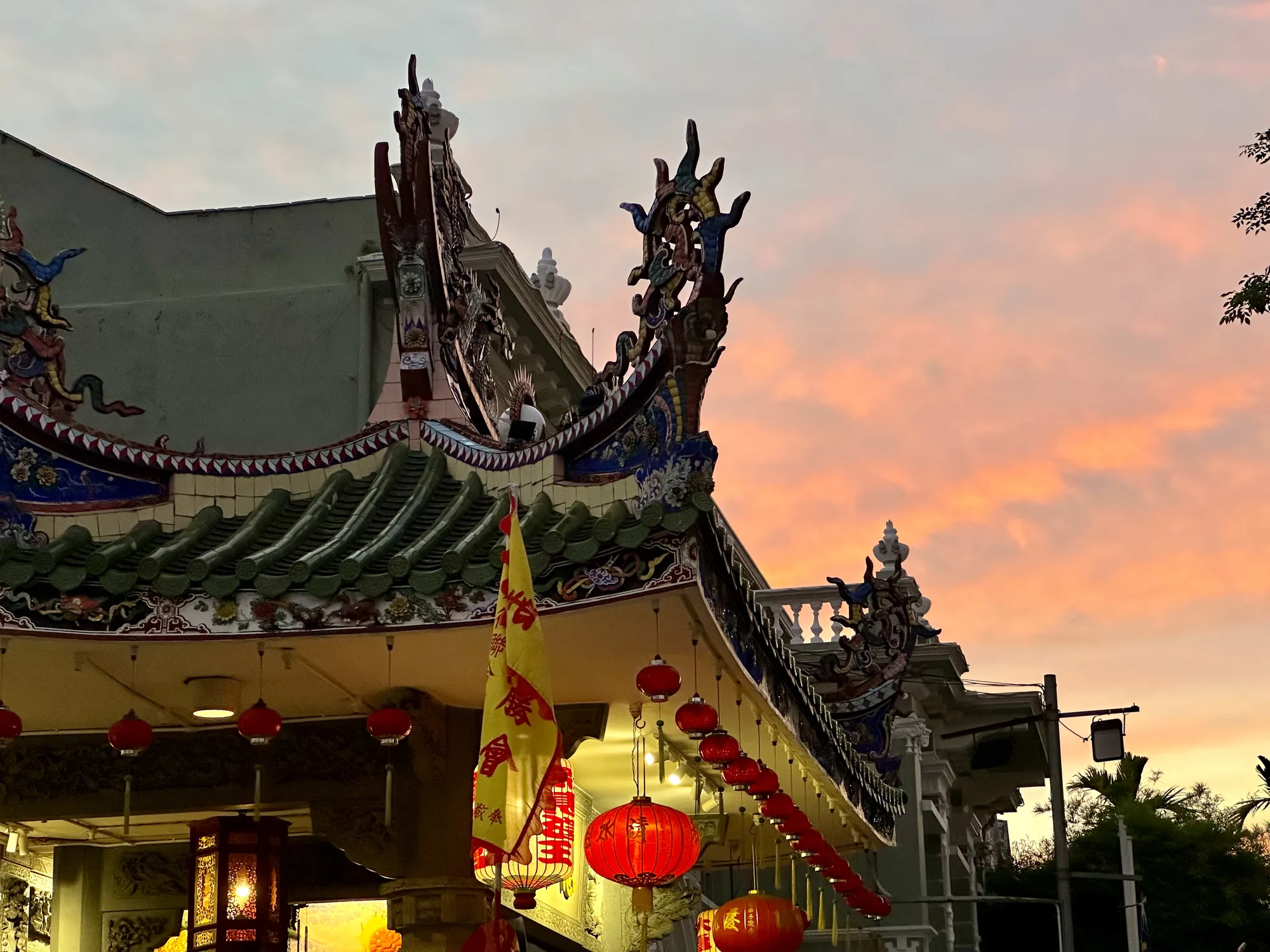 マレーシア、ペナンで撮影された写真です。夕焼け空を背景に、中国寺院の一部が写っています。寺院の屋根は緑色の瓦で覆われており、精巧な装飾が施されています。屋根の端には、龍や鳥などの彫刻が並んでいます。軒下には、赤い提灯が連なって吊るされており、夕暮れ時の柔らかな光を浴びて幻想的な雰囲気を醸し出しています。黄色い旗も立てられており、寺院の荘厳さを際立たせています。空はピンク色からオレンジ色へとグラデーションを描き、美しい夕焼けが寺院全体を包み込んでいます。