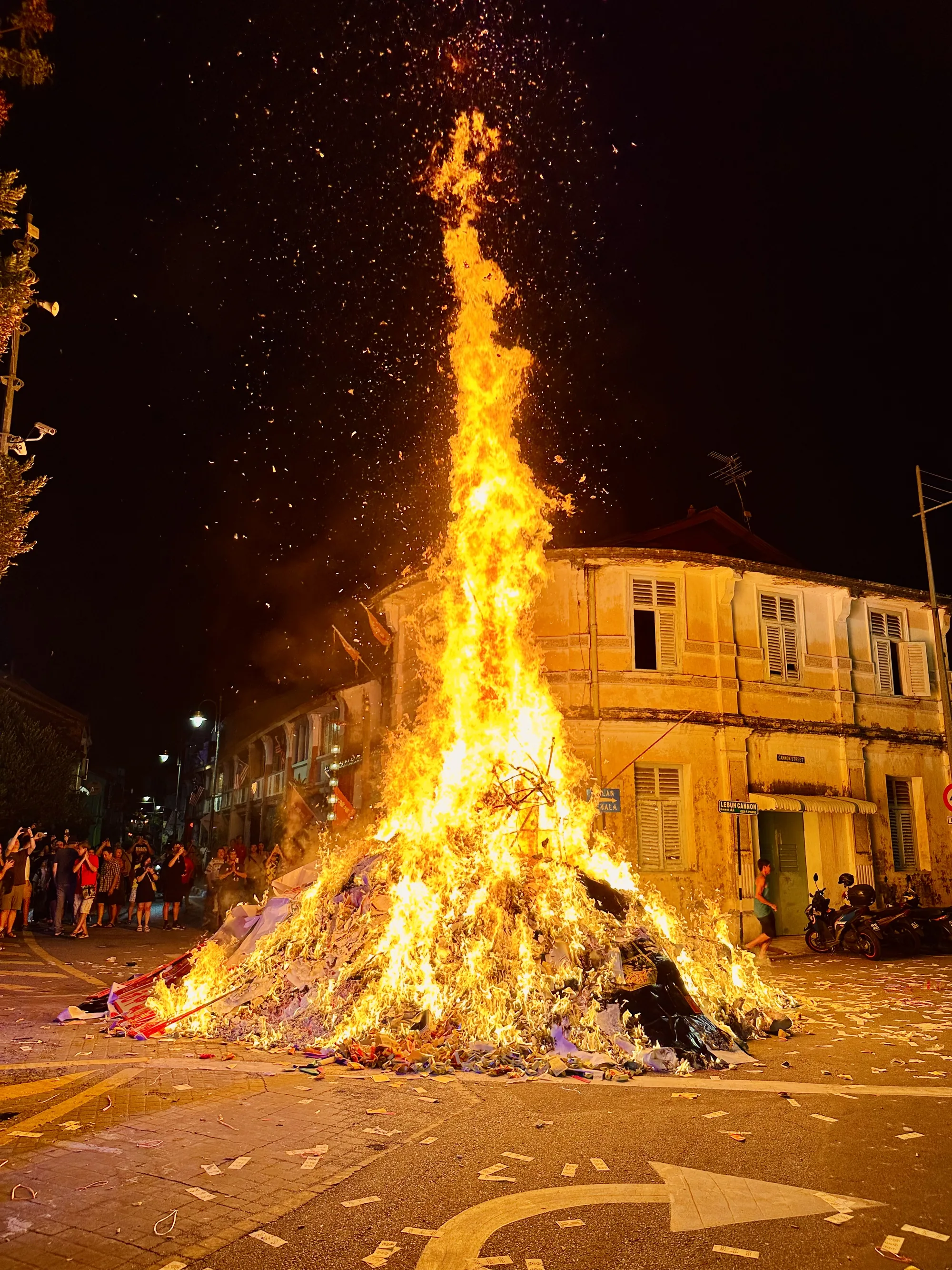 マレーシアのペナン島で撮影された夜景写真です。道路の中央に巨大な焚き火が燃え盛っており、炎は高く立ち上り、火の粉が夜空に舞い散っています。焚き火の周りには、それを囲んで見物する人々の姿が見えます。人々は焚き火の様子を写真やビデオに収めているようです。道路には、焚き火の灰や燃えカスのようなものが散らばっており、紙幣のようなものが落ちています。背景には、古い建物が並んでおり、一部の建物には窓が開け放たれています。建物の前にはバイクが数台駐車されています。写真全体は、夜の熱気と興奮に包まれたような雰囲気で、お祭りのようなイベントの様子を捉えています。