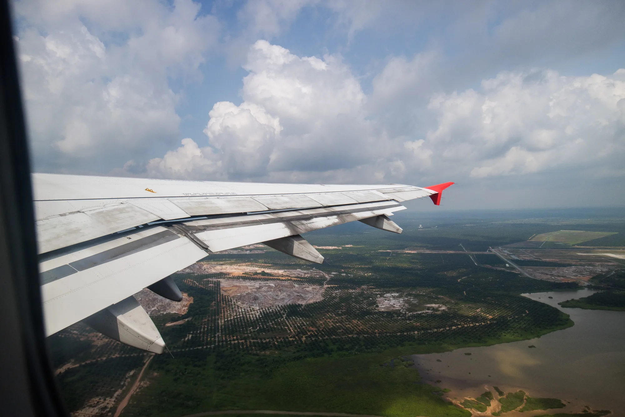 写真は、マレーシアのペナン上空を飛行する飛行機から撮影されたものです。画面左側には、飛行機の窓枠の一部が見えています。窓の外には、白い雲が浮かぶ青空が広がっています。写真の中央から下にかけては、飛行機の白い翼が写っており、翼端は赤色に塗られています。翼の下には、緑豊かな森林や水面が広がっており、その奥には道路や建物のような人工的な構造物も見えます。全体的に明るい光が差し込み、爽やかな雰囲気が感じられます。
