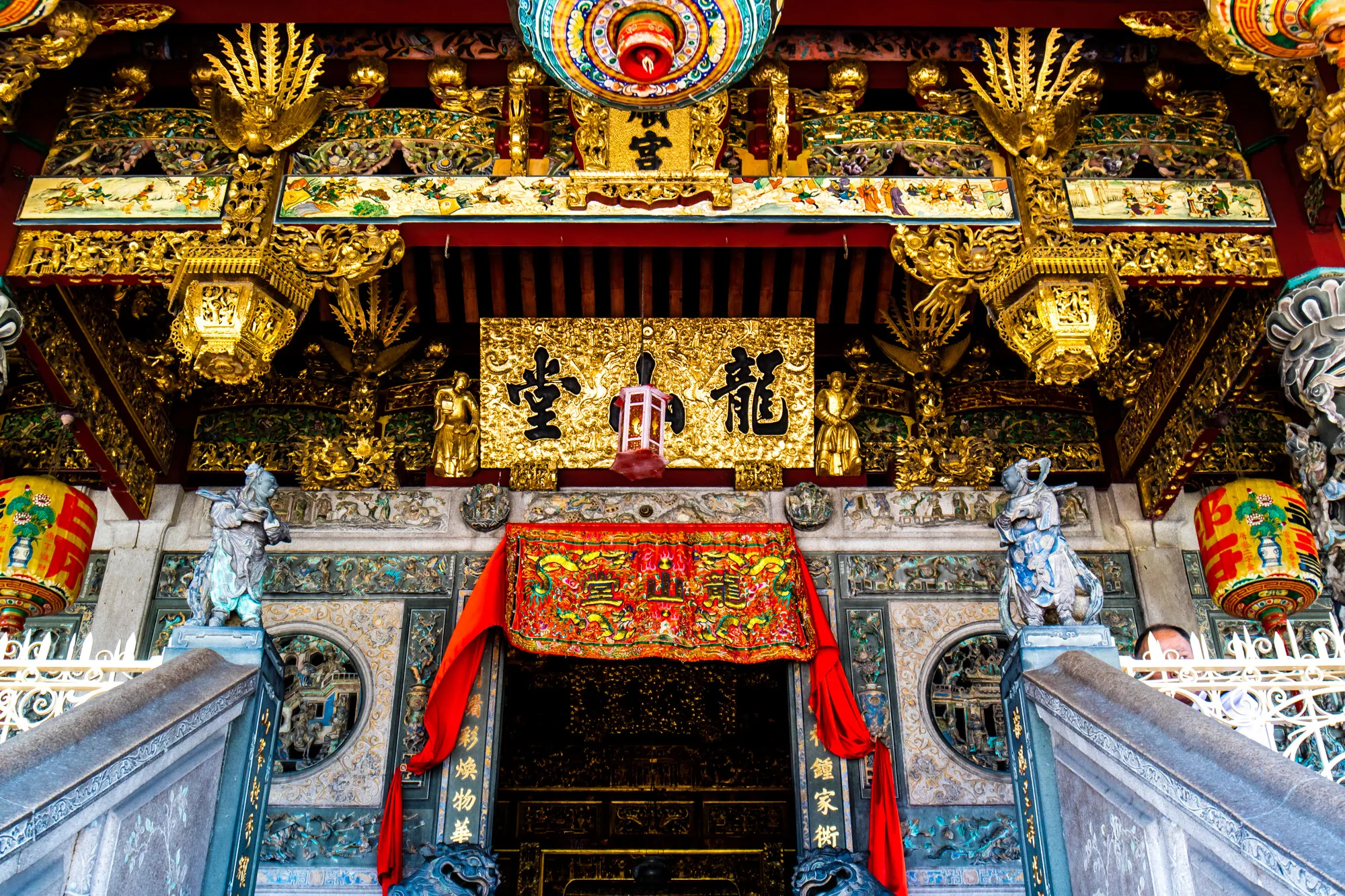 ペナンの寺院の入り口を正面から捉えた写真です。入り口は階段で少し高くなっており、石造りの階段の左右には狛犬のような像が鎮座しています。階段の上には、細やかな装飾が施された入口があり、中央には赤い垂れ幕がかかっています。入口の上部には、「龍山堂」と書かれた金色の額が掲げられています。額の左右には黄金の像が配置され、さらにその上には、鮮やかな装飾が施された天井が見えます。全体的に金色、赤色、青色などが多用されており、非常に華やかで荘厳な雰囲気です。
