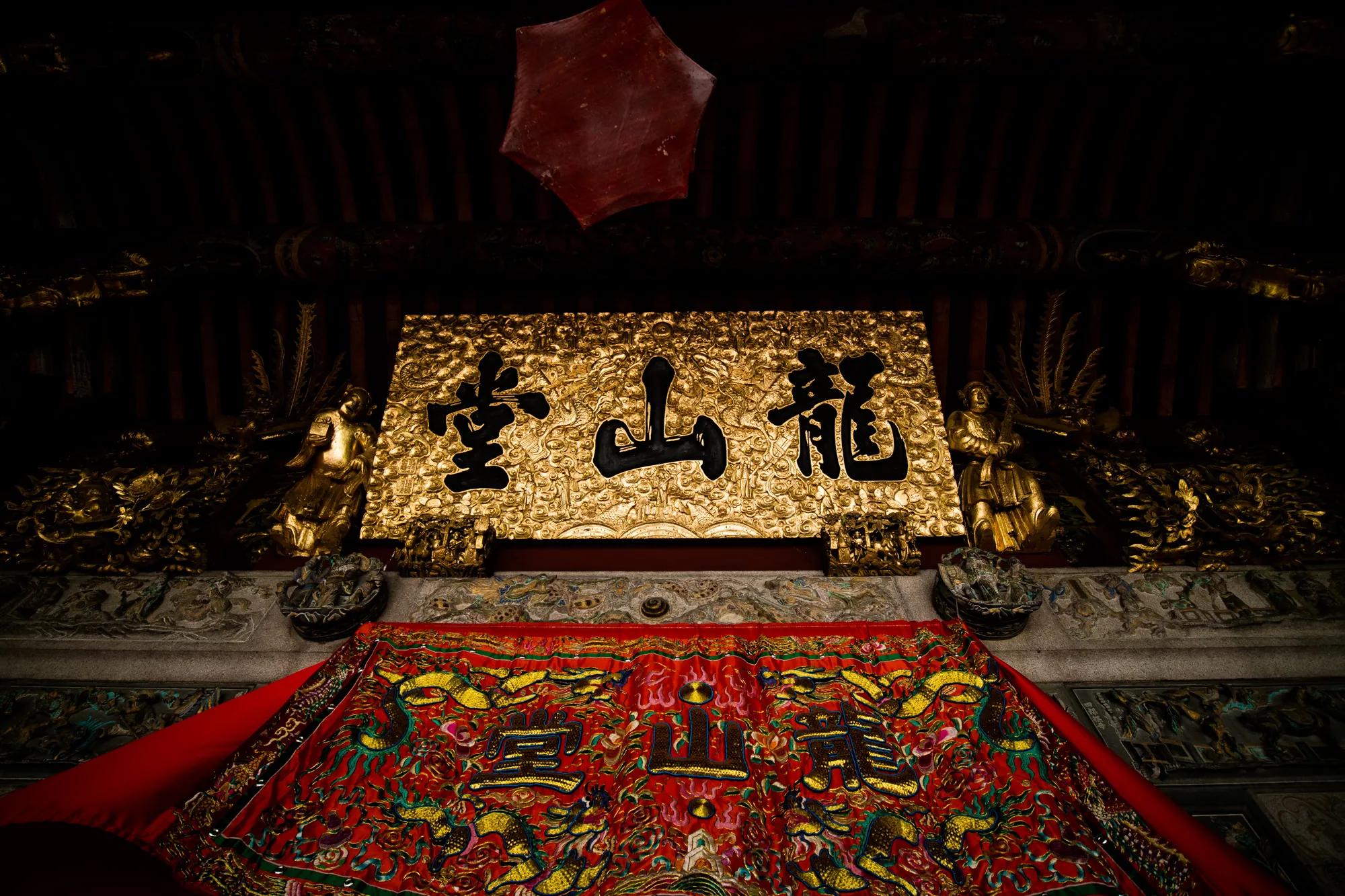 マレーシアのペナンで撮影された、寺院の内部を捉えた写真です。全体的に暗く、照明によって金色や赤色が強調された厳かな雰囲気を醸し出しています。  写真の中心には、金色の装飾が施された看板があり、黒色の文字で「龍山堂」と書かれています。看板の両側には、金色の彫像が配置されています。看板の下には、赤色を基調とした鮮やかな刺繍が施された布が掛けられています。この布にも、金色の文字で「龍山堂」の文字が見られます。  天井には、装飾が施されており、赤い六角形の装飾品が吊るされています。写真全体から、伝統的な中国様式の建築様式と、信仰の深さが感じられます。