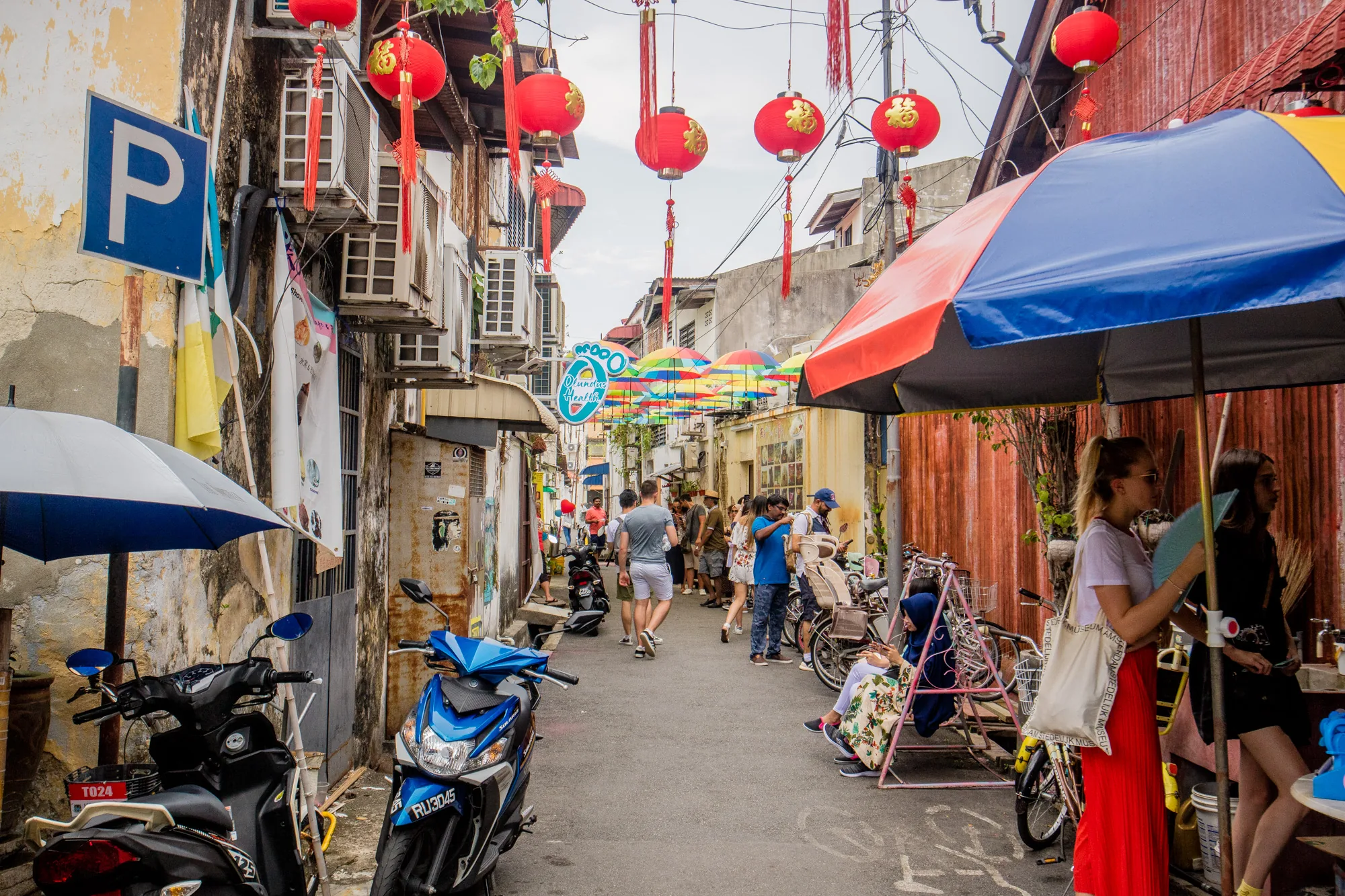 写真はマレーシアのペナンで撮影されたもので、賑やかな路地の様子を捉えています。路地の上には赤い提灯が吊るされ、さらに奥にはカラフルな傘が連なっています。路地の両側には古い建物が立ち並び、壁にはエアコンの室外機が多数設置されています。路地には様々な人々が行き交い、観光客や地元の人々が混ざり合っているようです。路地の脇にはバイクや自転車が停められ、生活感が感じられます。写真全体は活気にあふれ、ペナンの独特な雰囲気を伝えています。路地の入口には「P」の駐車場標識があり、路地の手前には青い傘と黒と青のバイクが写っています。