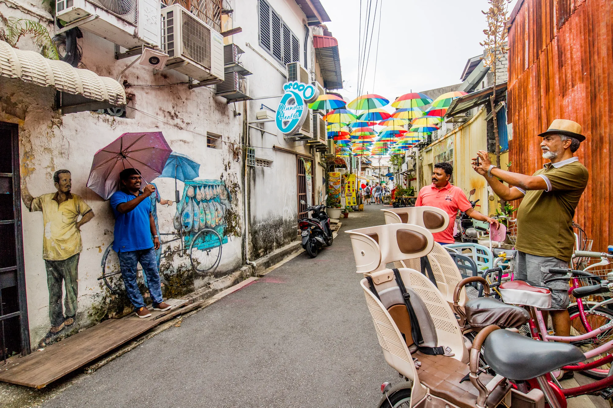 写真はマレーシアのペナンで撮影されたもので、賑やかな街の様子が捉えられています。細い路地には、カラフルな傘が空を覆うように飾られており、観光名所となっているようです。路地の壁には、人物や魚の絵が描かれており、独特の雰囲気を醸し出しています。写真には、壁の絵の前でポーズをとる男性や、自転車に寄りかかってスマートフォンで写真を撮る男性など、地元の人の姿も見られます。古い建物と新しい装飾が混ざり合った、ペナンの魅力的な雰囲気が伝わってくる一枚です。路地の奥には、観光客らしき人々も写っており、活気のある様子が伺えます。
