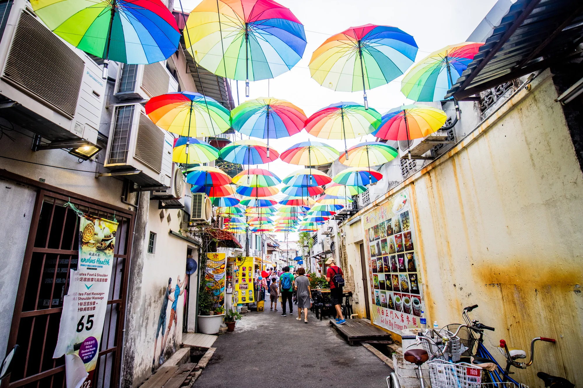 マレーシアのペナンで撮影された写真です。細い路地を撮影したもので、路地の頭上にはたくさんのカラフルな傘が吊り下げられています。傘は赤、青、緑、黄色など、さまざまな色で彩られています。路地の両側には建物が並び、エアコンの室外機がたくさん設置されています。壁には看板やポスターが貼られています。路地には観光客らしき人々が歩いており、賑やかな雰囲気です。路地の奥に向かって遠近感が強調されています。 自転車も止まっており、生活感も感じられます。全体的に明るく、カラフルで楽しい雰囲気の写真です。