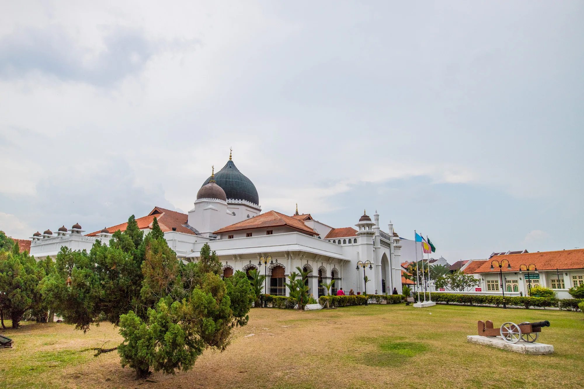 マレーシアのペナンで撮影された写真には、美しいモスクが写っています。建物は白を基調とし、オレンジ色の屋根瓦と黒いドームが特徴的です。モスクの周りには緑豊かな芝生が広がり、手前にはいくつかの木々が植えられています。芝生の一角には大砲の模型が置かれています。建物の背景には、いくつかの旗がはためいており、歴史的な雰囲気を醸し出しています。空は曇り空で、全体的に穏やかで静かな印象を受けます。