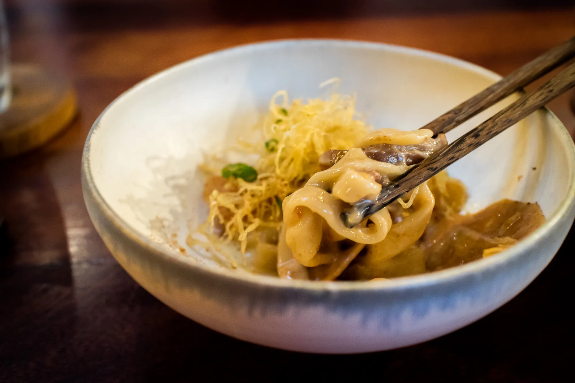 マレーシアのペナンで撮影された写真です。写真は、木のテーブルの上に置かれた白いボウルに入った麺料理をクローズアップで捉えています。料理は、もちもちとした麺、きのこのような具材、そして揚げ麺のようなものが添えられています。箸で麺を持ち上げており、その表面にはソースがかかっているのが見えます。全体的に美味しそうで、食欲をそそるような雰囲気です。照明はやや暗めで、料理にスポットライトが当たっているように見えます。背景には、ぼやけたグラスのようなものが見えます。