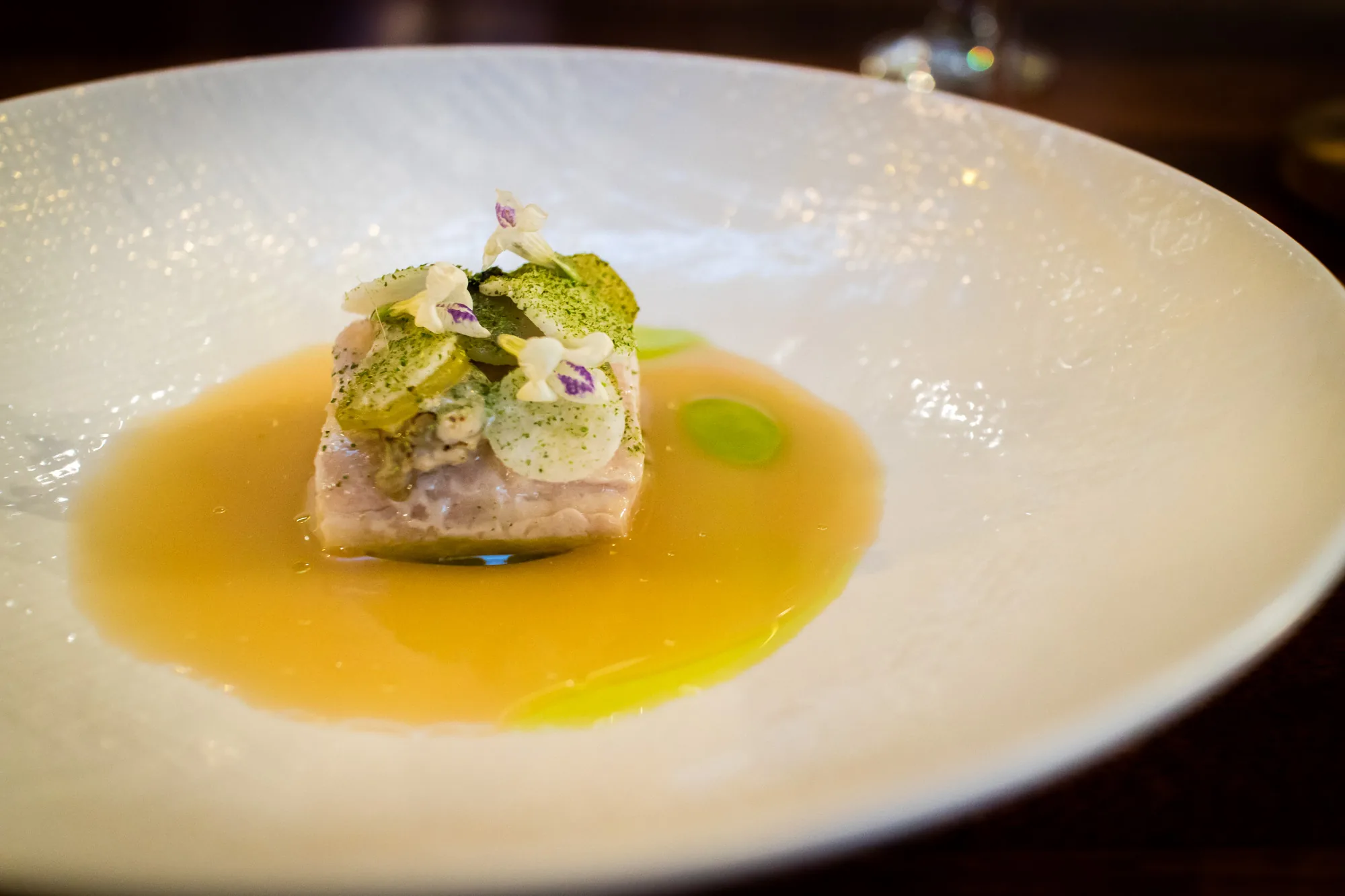 マレーシアのペナンで撮影されたと思われる写真には、白い皿に盛り付けられた料理が写っています。料理の中心には、角切りにされた魚の切り身があり、上には緑色の粉末や白いスライス、そして小さな白い花が添えられています。魚の周りには、鮮やかなオレンジ色のソースが広がっています。皿の縁は波打つようなデザインで、高級感があります。奥には、グラスの脚のようなものがぼんやりと写っており、レストランの一場面であることが伺えます。全体的に、上品で繊細な印象を与える料理の写真です。