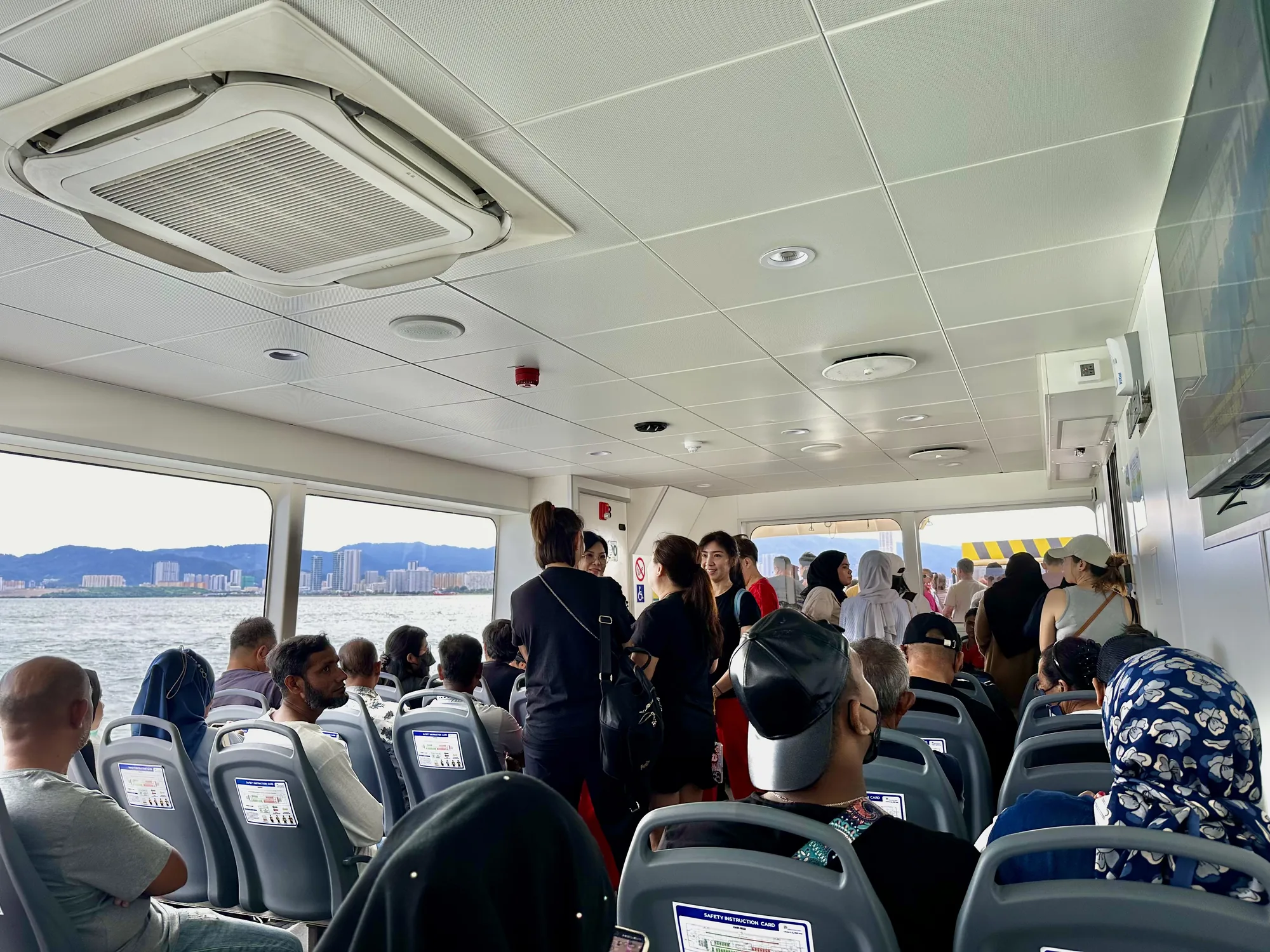 写真はマレーシアのペナンで撮影されたフェリーの船内の様子を捉えています。多くの人々が座席に座ったり、立って話したりしており、活気のある雰囲気です。窓からは海と、遠くにペナンの都市の建物が見えます。船内は白を基調としており、天井にはエアコンが設置されています。座席の背もたれには安全に関する説明書が貼られています。多様な服装の人々がおり、文化的な多様性も感じられます。