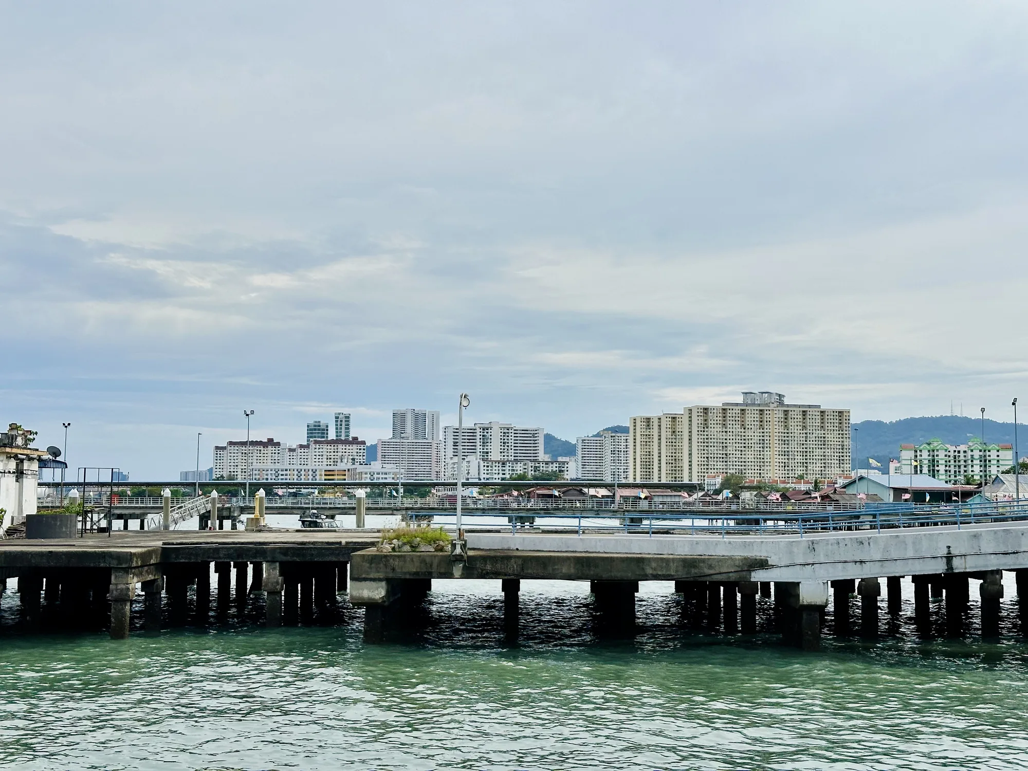 マレーシア、ペナン島で撮影された写真です。前景には、海に突き出したコンクリート製の桟橋が写っています。桟橋は、多数の柱によって支えられており、柱の影が海面に落ちています。海面は穏やかで、わずかに波立っており、緑がかった色をしています。  背景には、ペナン島の都市景観が広がっています。高層ビルやアパートメントが立ち並び、その背後には低い山々が見えます。建物は白やベージュの色調で、整然と並んでいます。一部の建物の間には、マレーシアの国旗が掲げられているのが見えます。桟橋と建物の間には、細い通路のようなものが走っており、その上には屋根付きの建物が並んでいるのが見えます。  空は曇っており、全体的に落ち着いた、やや霞んだ雰囲気です。全体として、この写真はペナン島の港の日常的な風景を捉えており、現代的な都市と伝統的な要素が混在している様子が伺えます。